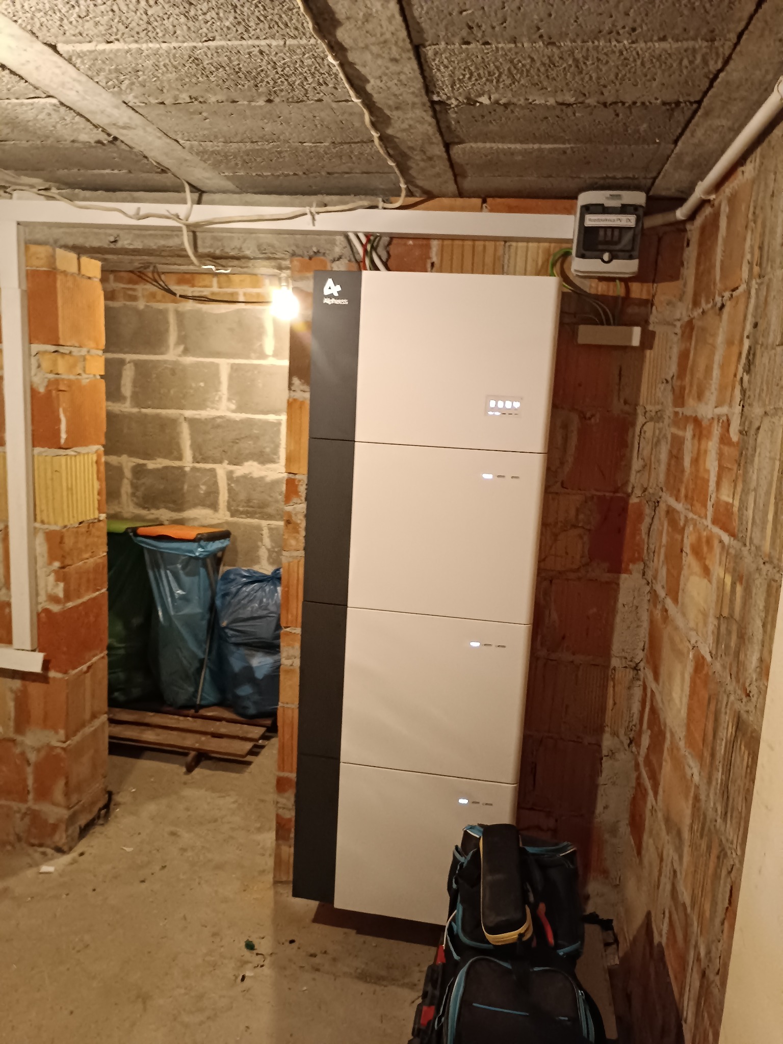 Biała, modułowa bateria energii z czarnym panelem bocznym w surowym wnętrzu. Widoczne kable, cegły, narzędzia. Minimalistyczny design, potencjalnie instalacja fotowoltaiczna.