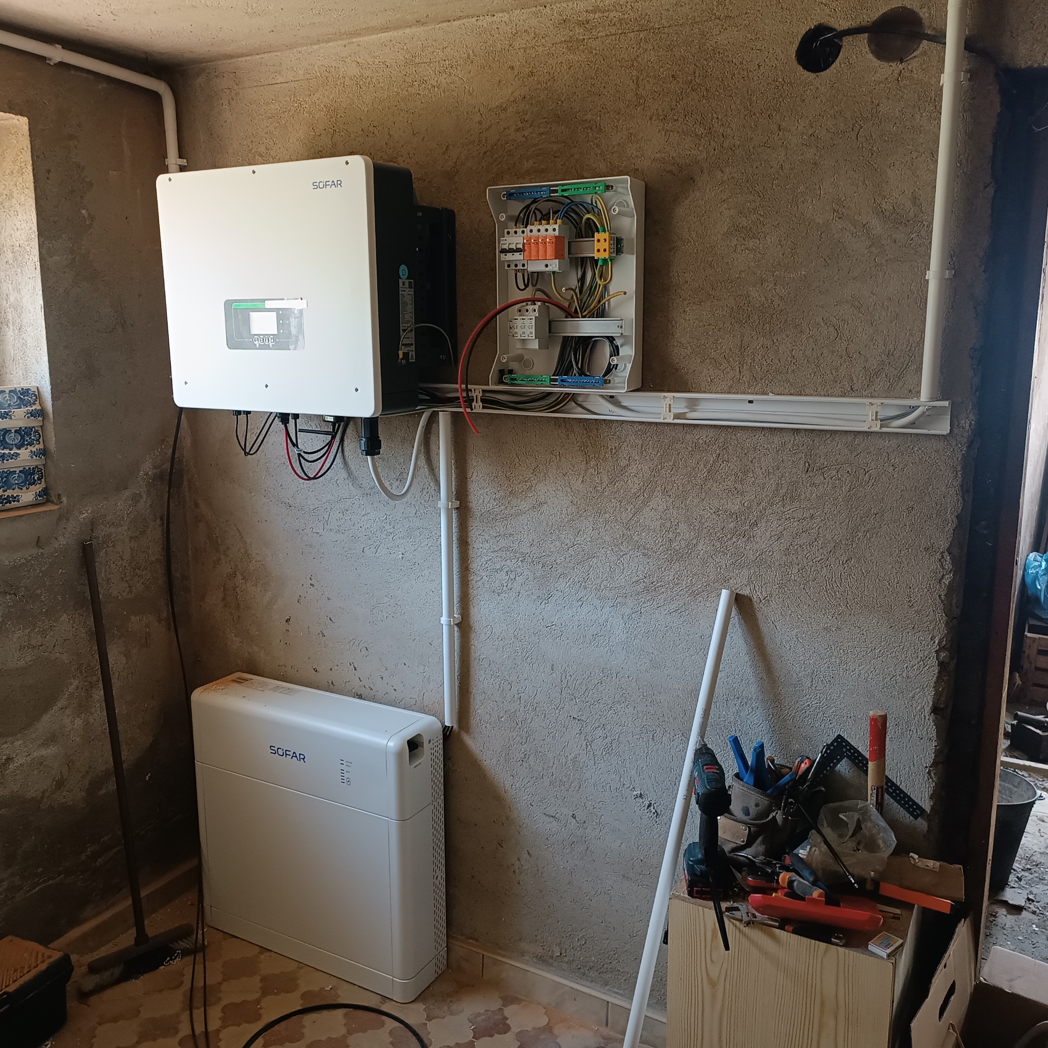 Instalacja paneli fotowoltaicznych SOFAR: falownik, akumulator i skrzynka z bezpiecznikami na surowej ścianie. Widoczne narzędzia i instalacja elektryczna.