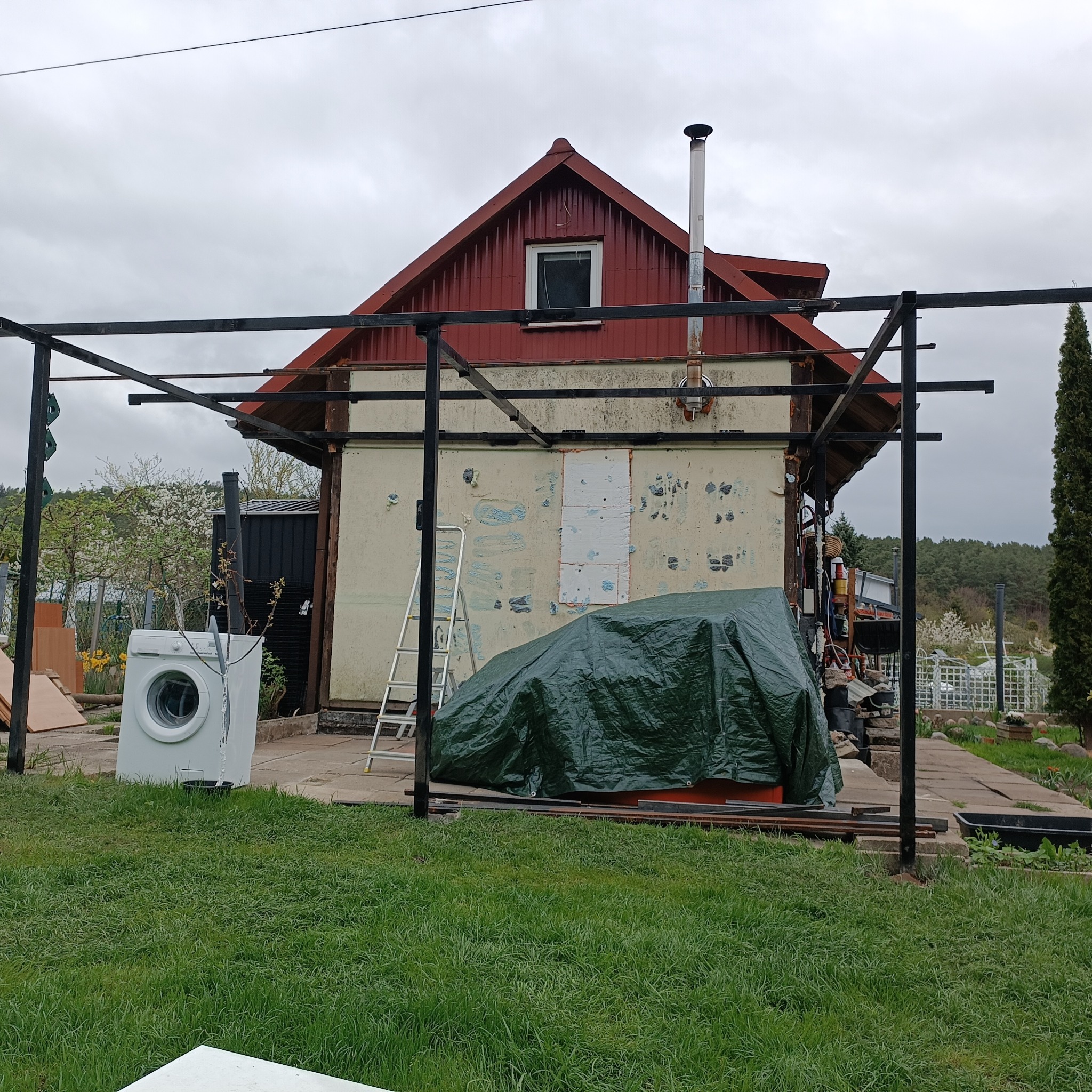 Dom w trakcie remontu elewacji, z widoczną drabiną, przykrytym sprzętem plandeką, pralką na zewnątrz i konstrukcją z metalowych profili. Tło: drzewa, pochmurne niebo.