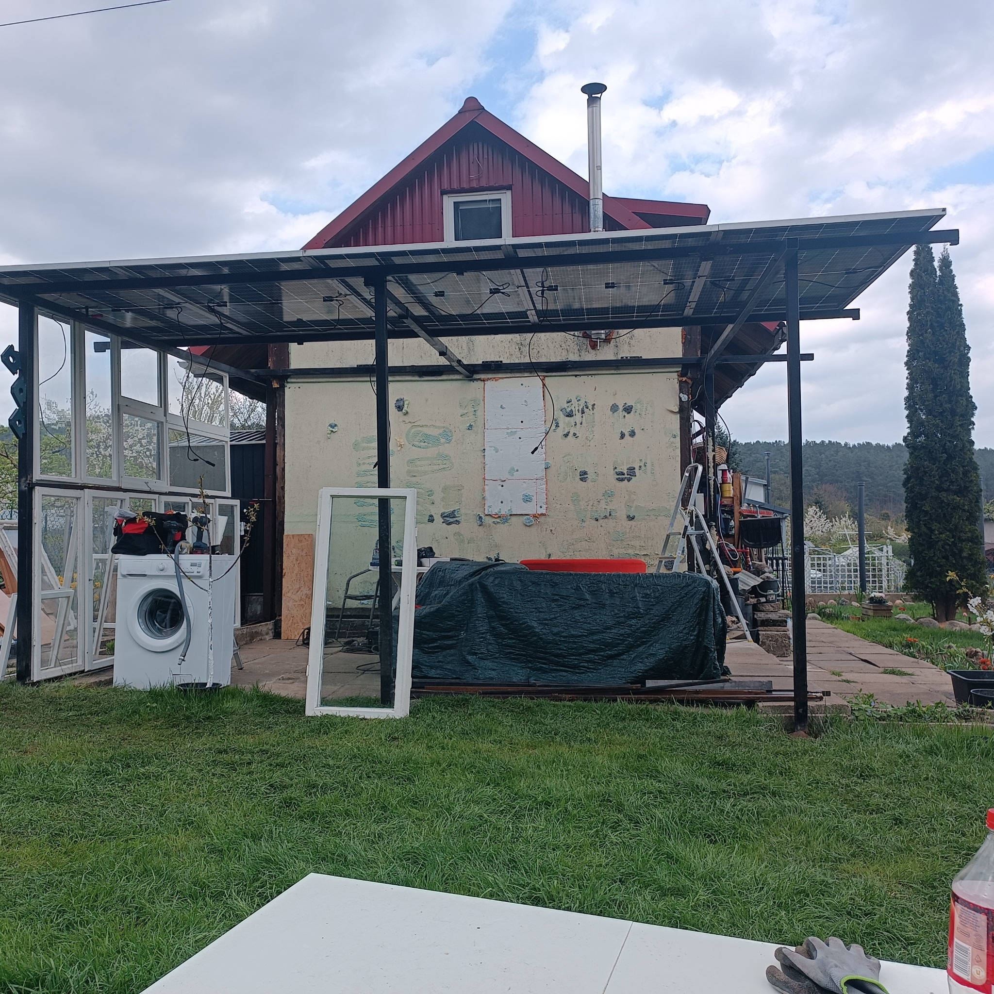 Dom z panelami słonecznymi w trakcie instalacji. Widoczne rusztowanie, pralka, okna i narzędzia. Nieukończony montaż, bałagan na posesji.