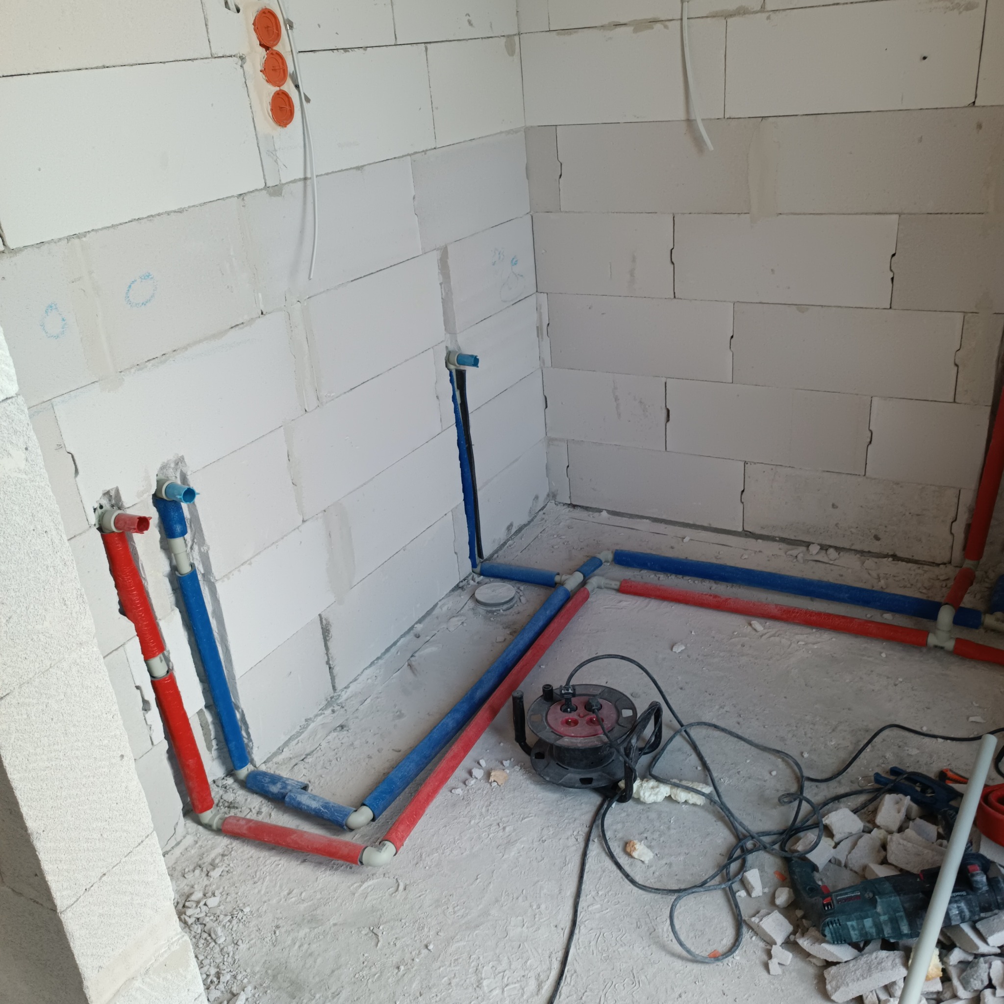 Instalacja wodna w surowym wnętrzu: czerwone i niebieskie rury z tworzywa sztucznego prowadzone wzdłuż ścian z białych bloczków. Widoczne narzędzia i przedłużacz.