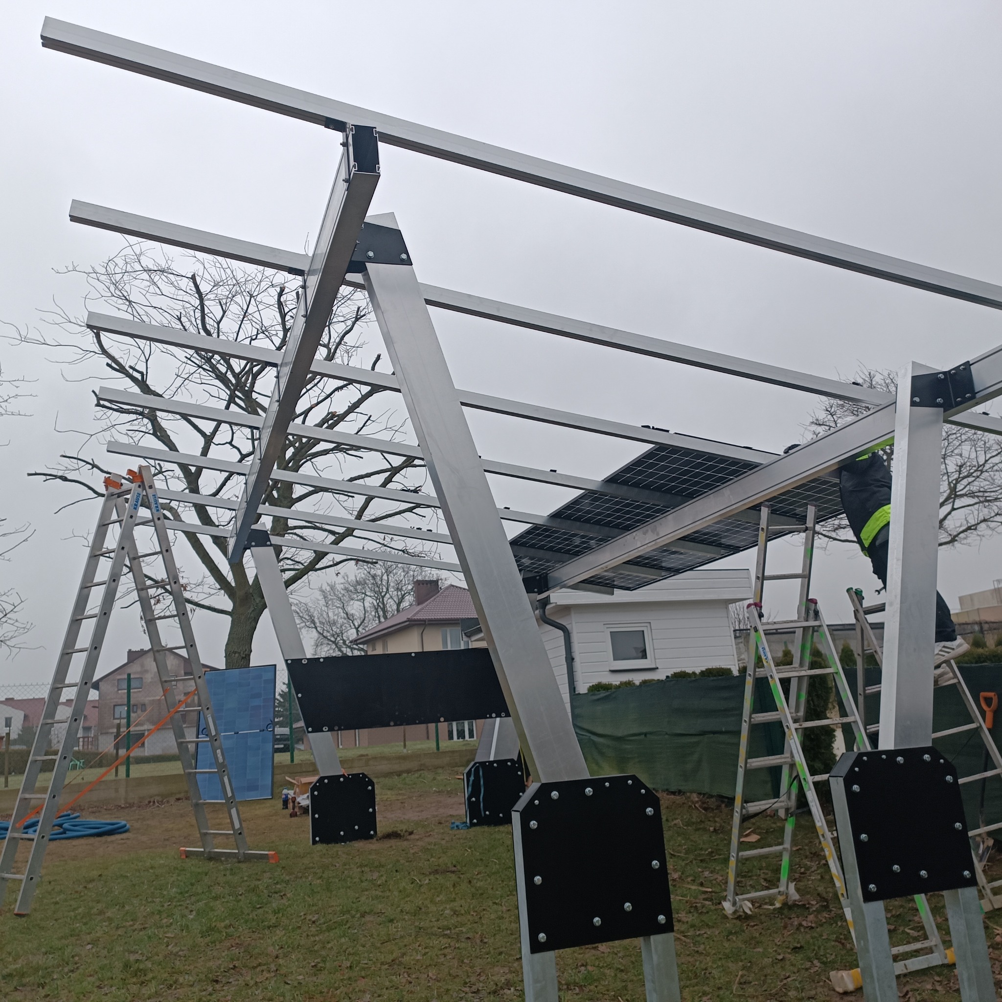 Montaż konstrukcji pod panele fotowoltaiczne na zewnątrz, drabiny, częściowo zamontowane panele, pochmurne niebo w tle, widoczne domy jednorodzinne.