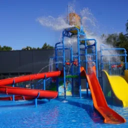 Kompleks zjeżdżalni wodnych w jaskrawych kolorach (czerwony, żółty, pomarańczowy) z wiadrem wylewającym wodę, umieszczony w basenie z błękitną wodą.