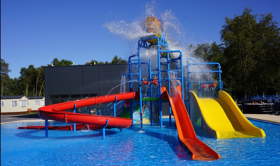 Kompleks zjeżdżalni wodnych w jaskrawych kolorach (czerwony, żółty, pomarańczowy) z wiadrem wylewającym wodę, umieszczony w basenie z błękitną wodą.