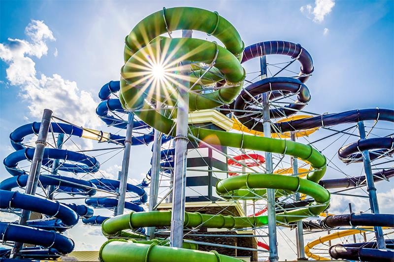 Kompleks zjeżdżalni wodnych w parku rozrywki, z wieżą w tle i promieniami słońca przebijającymi przez konstrukcję. Kolorowe rury tworzą spiralne wzory na tle błękitnego nieba.