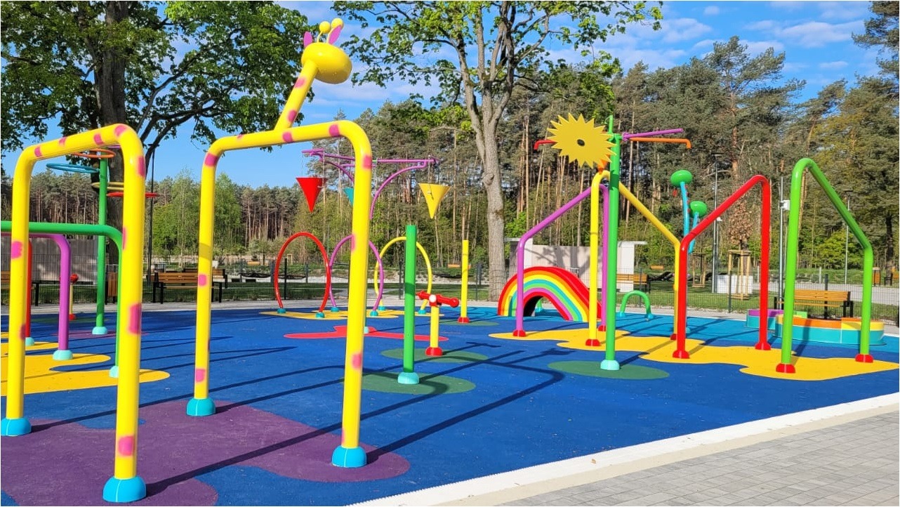 Nowoczesny, kolorowy plac zabaw wodny z motywem żyrafy, tęczy i słońca na niebieskiej, gumowej nawierzchni, otoczony drzewami i ławkami. Widok za dnia.