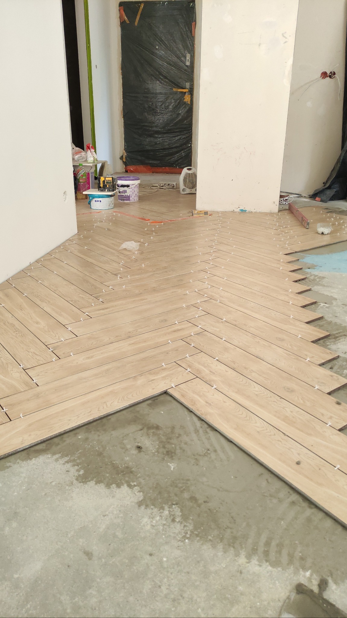 Układanie jasnych paneli podłogowych w jodełkę. Widoczne krzyżyki dystansowe, poziomica i przygotowane materiały. Prace wykończeniowe w trakcie realizacji.