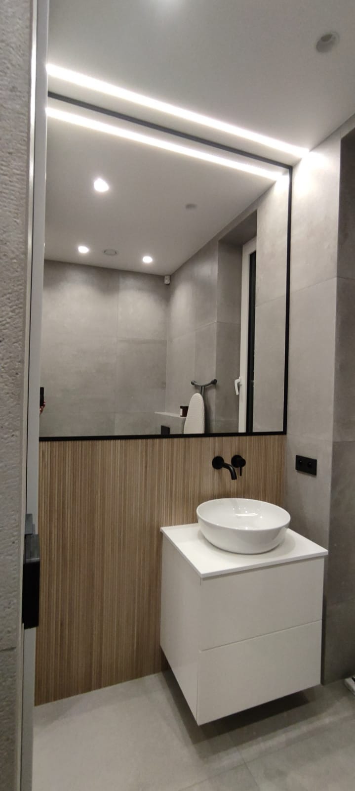 Nowoczesna łazienka: biała umywalka nablatowa, czarna bateria, lustro z podświetleniem LED. Ściana wykończona płytkami i drewnianymi panelami. Minimalistyczny design.