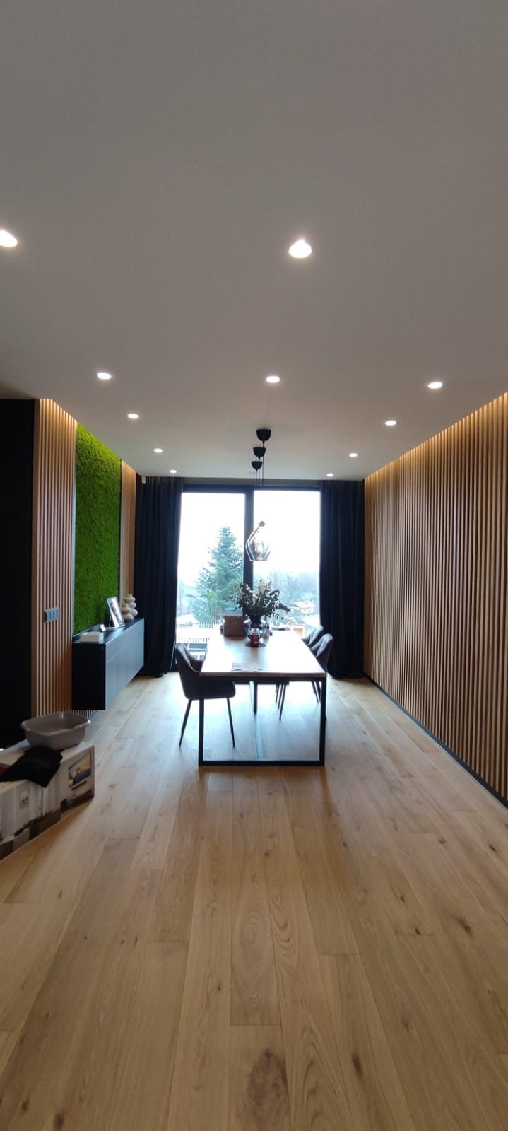 Jasny parkiet w przestronnym wnętrzu z drewnianymi lamelami na ścianach i zielonym panelem z mchu. Stół z krzesłami i widok na zewnątrz przez duże okno.