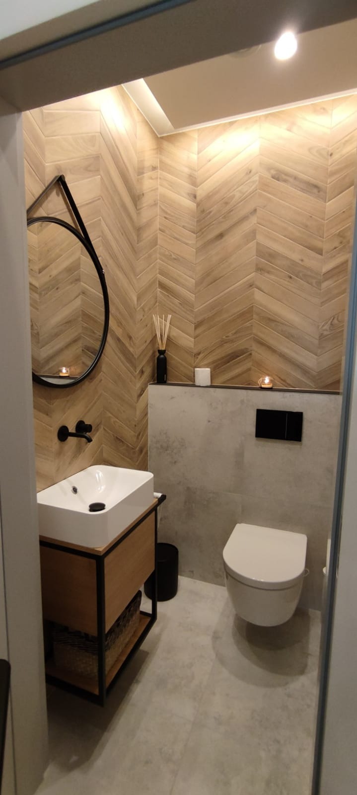 Nowoczesna łazienka z drewnopodobnymi płytkami ułożonymi w jodełkę, okrągłe lustro i minimalistyczna umywalka na szafce z czarną ramą.