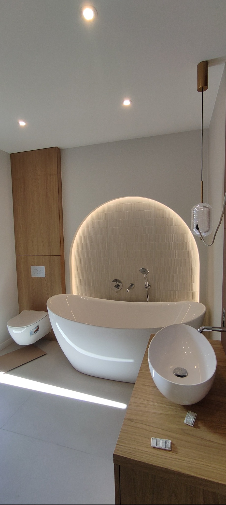 Nowoczesna łazienka z wolnostojącą wanną, umywalką nablatową i podświetlaną ścianą z mozaiki. Minimalistyczny design z drewnianymi akcentami i subtelnym oświetleniem.
