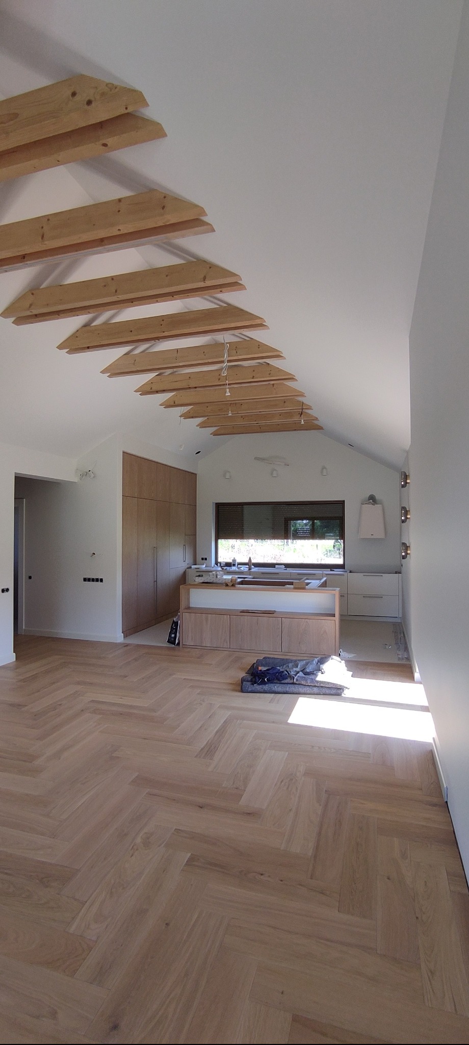 Jasne parkiety w jodełkę, drewniane belki stropowe i zabudowa meblowa w odcieniach drewna w przestronnym wnętrzu z oknem. Widok na fragment kuchni.