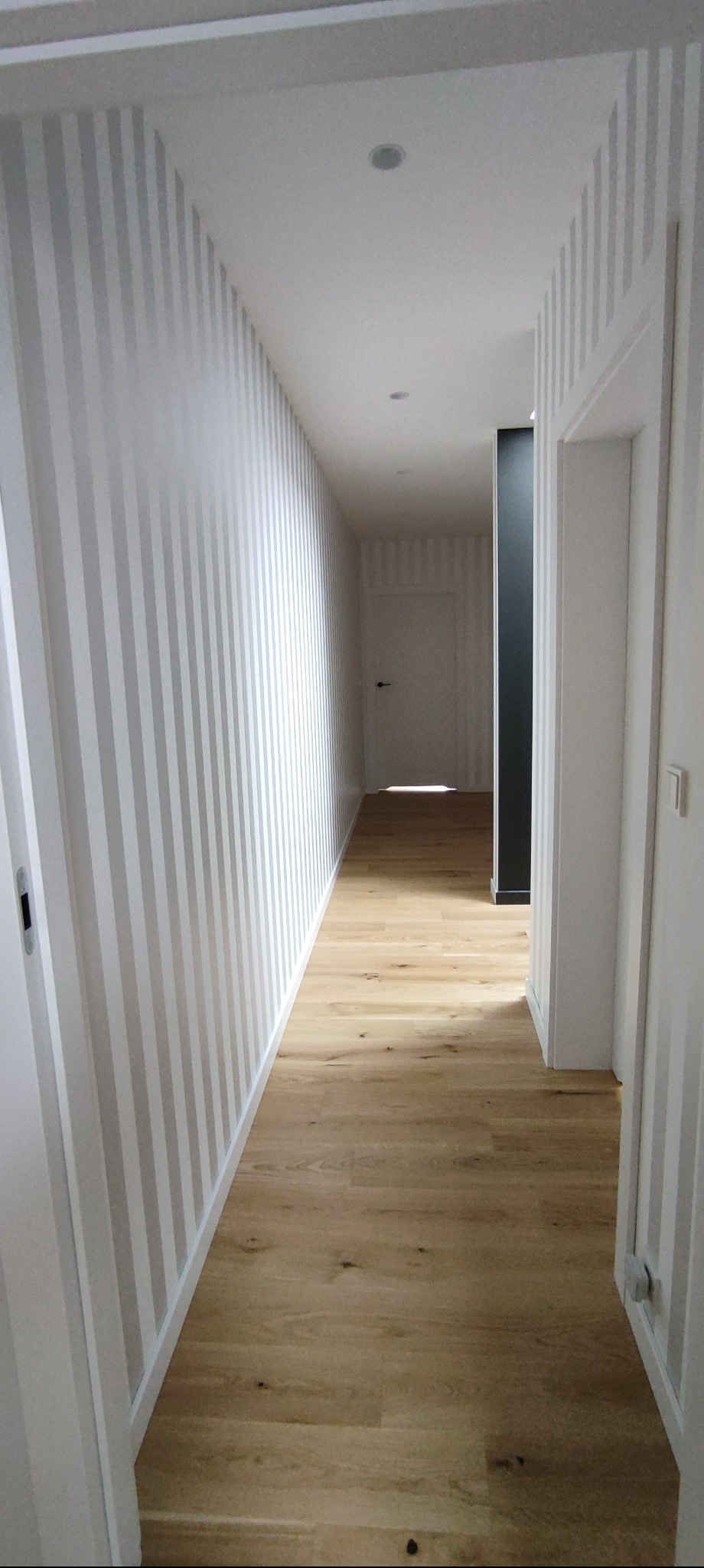 Wykończony korytarz z drewnianą podłogą i ścianami w biało-szare pasy. Widoczne drzwi i oświetlenie punktowe. Minimalistyczny design.