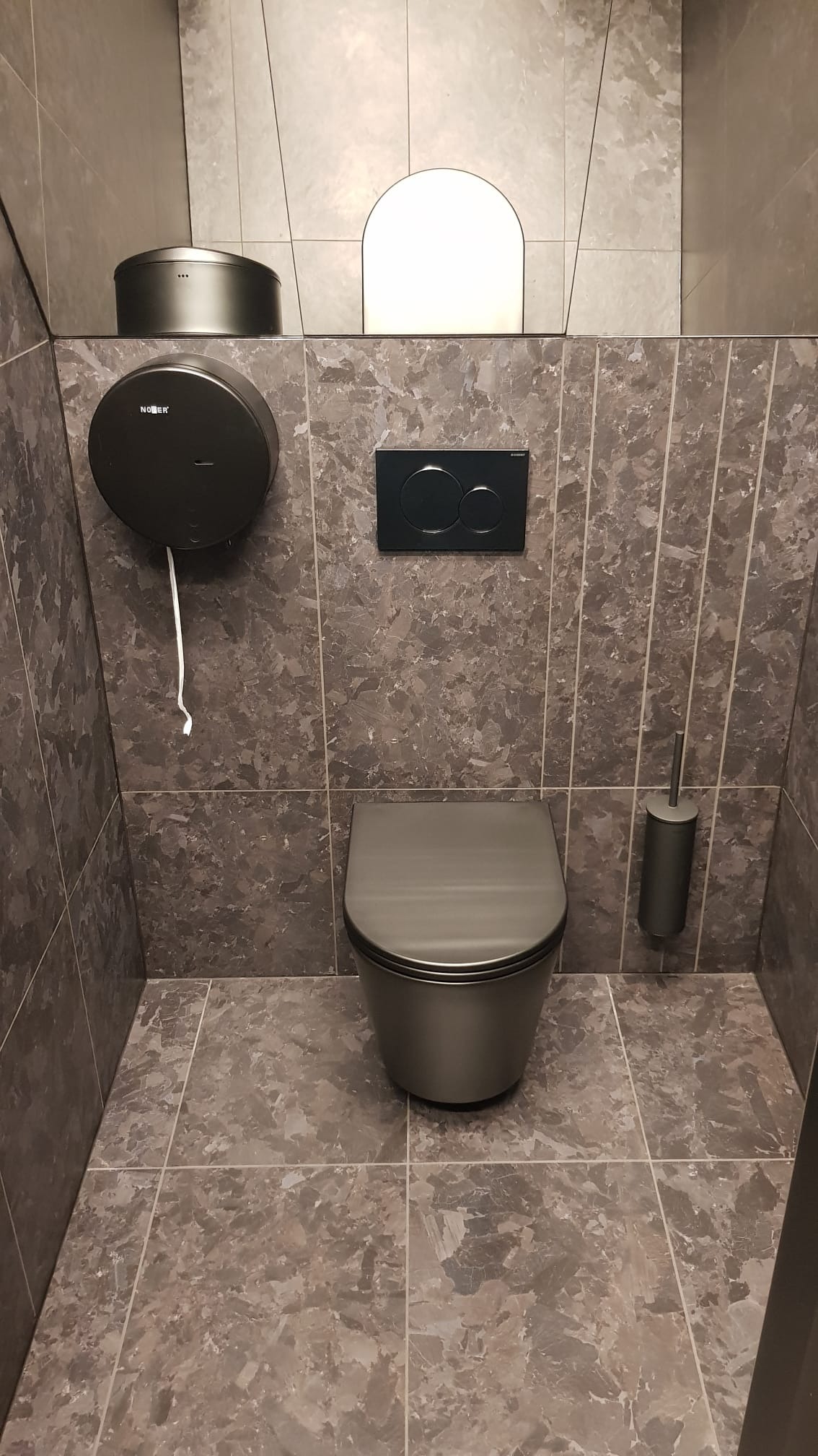 Nowoczesna toaleta w odcieniach szarości z marmurkową glazurą, wiszącą muszlą, czarnym przyciskiem spłukującym i pojemnikiem na papier toaletowy.