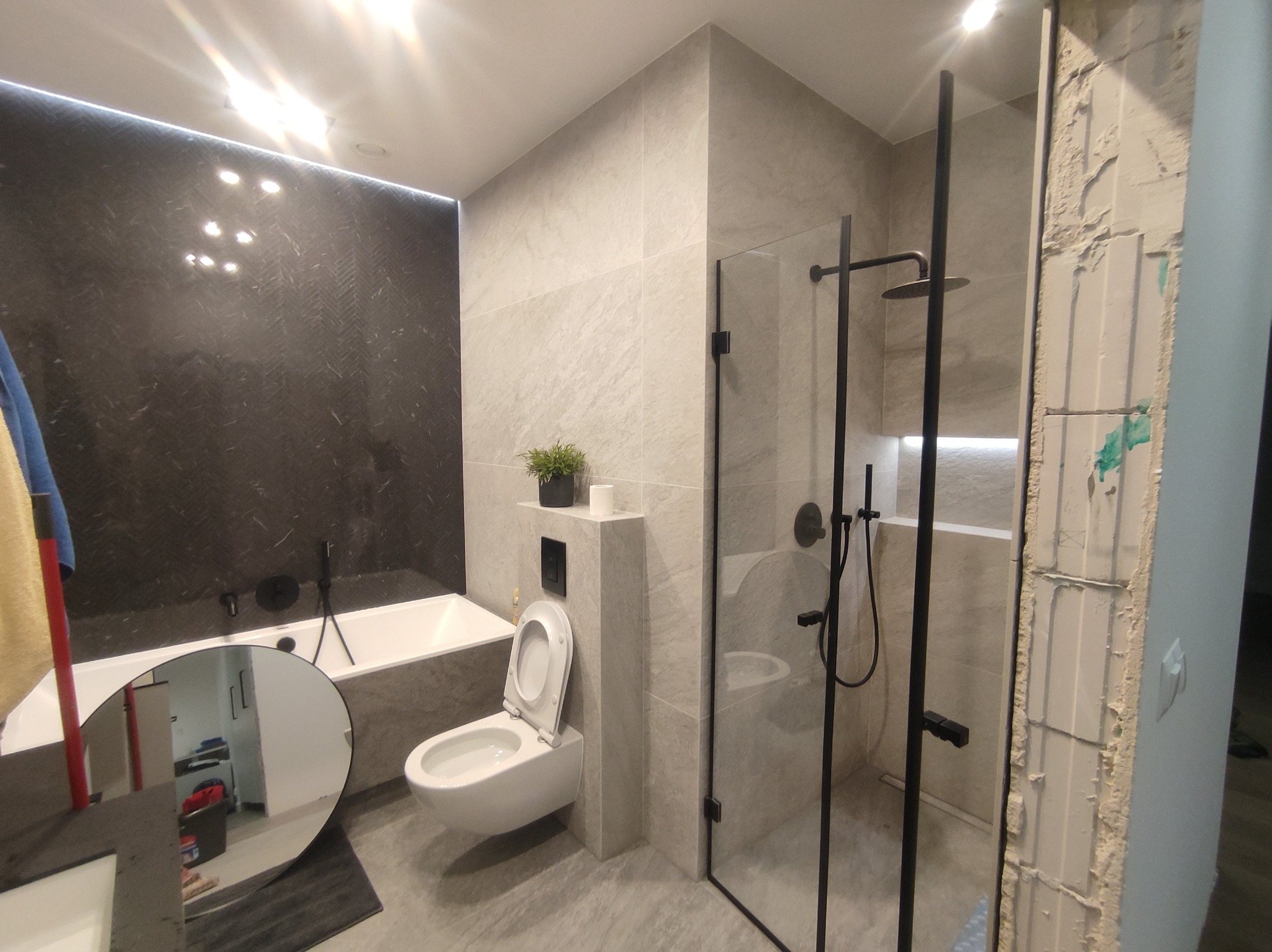 Nowoczesna łazienka w szarościach z czarną armaturą: wanna, toaleta podwieszana i kabina prysznicowa typu walk-in. Fragment ściany przed wykończeniem.