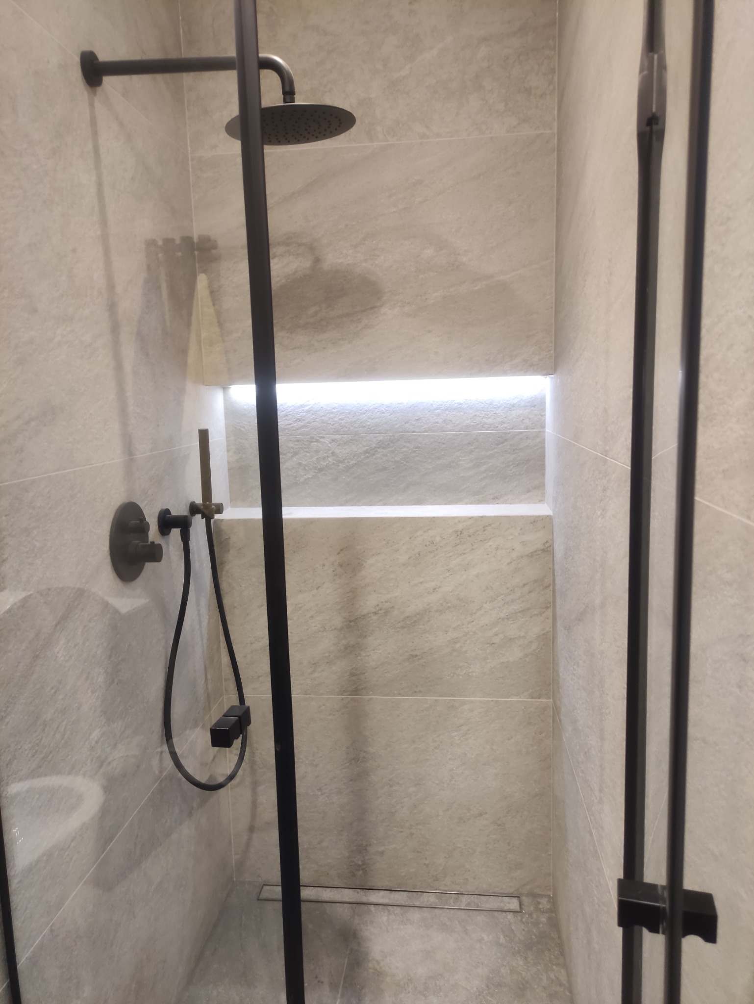 Nowoczesna kabina prysznicowa z czarną armaturą i wnęką z podświetleniem LED, wykończona płytkami imitującymi kamień w odcieniach szarości.