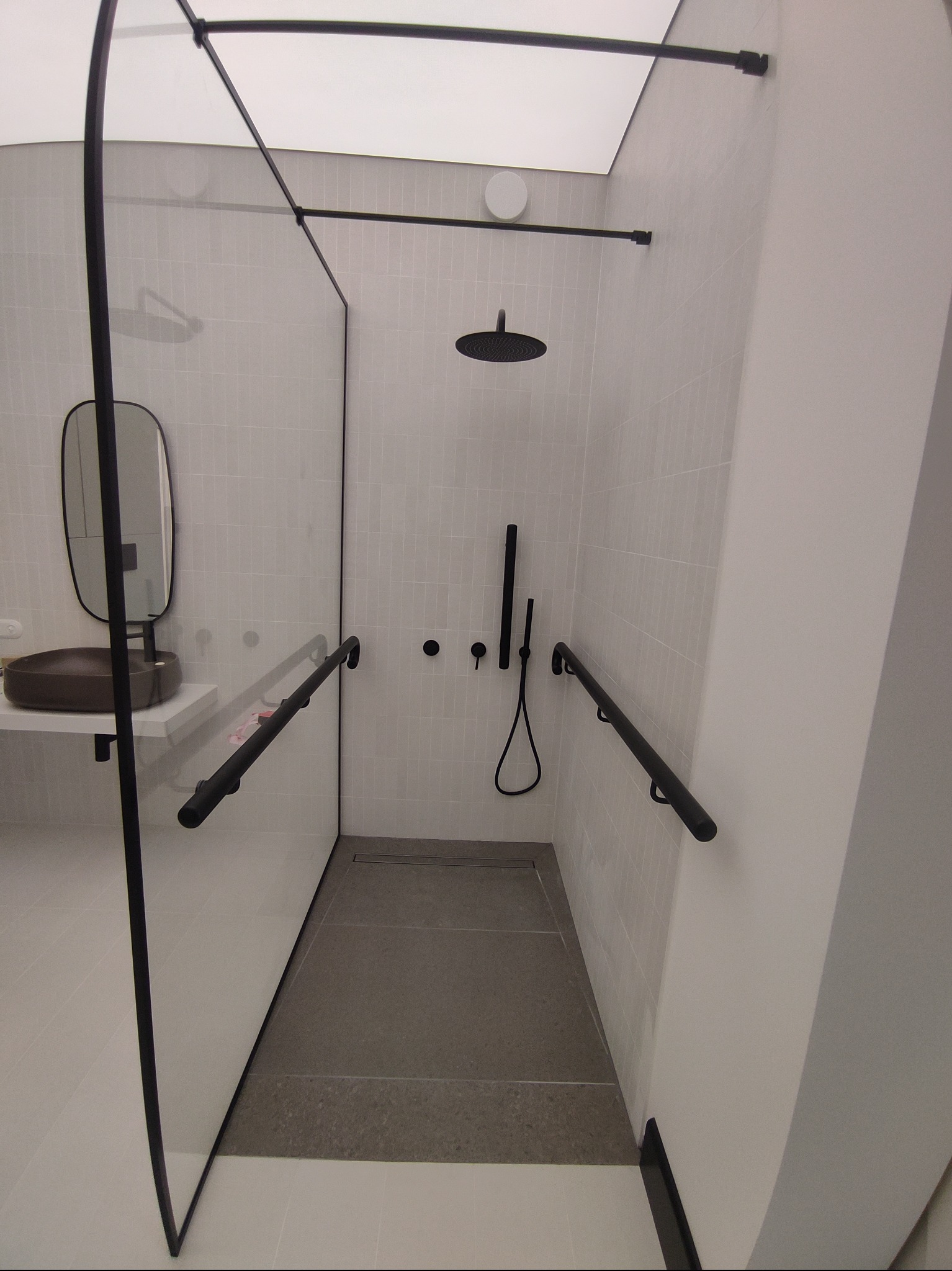 Nowoczesna łazienka z kabiną prysznicową typu walk-in, szare płytki, czarne detale armatury i minimalistyczna umywalka z lustrem.