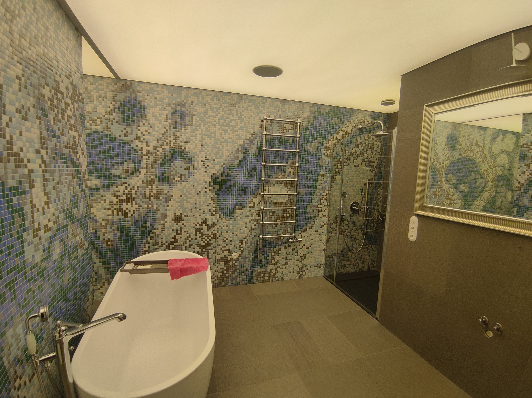 Nowoczesna łazienka z mozaikową ścianą, wolnostojącą wanną i kabiną prysznicową. Grzejnik drabinkowy i lustro w ozdobnej ramie dopełniają aranżację.