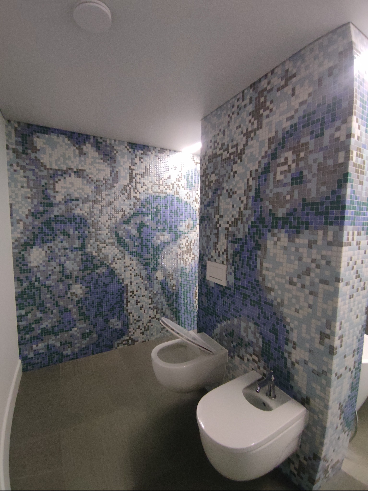 Mozaika w odcieniach błękitu i szarości zdobi ściany wykończonej łazienki z białą armaturą. Uwagę przykuwa wzór przypominający piksele, tworzący unikalny efekt wizualny.