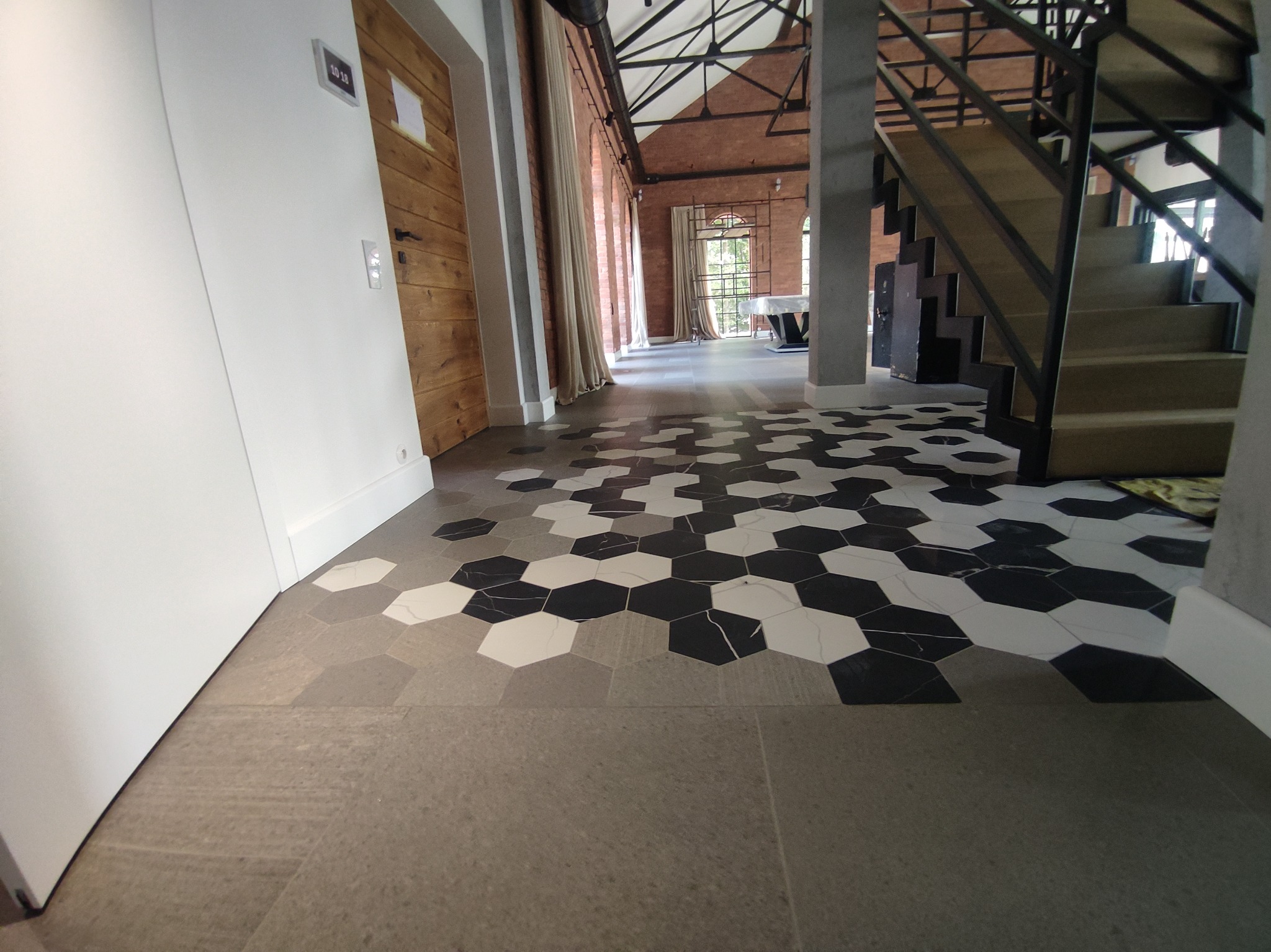 Industrialny hol z heksagonalną posadzką w odcieniach szarości, bieli i czerni, prowadzącą do schodów i przeszklonej przestrzeni. Widoczna drewniana stolarka drzwi i surowa cegła.
