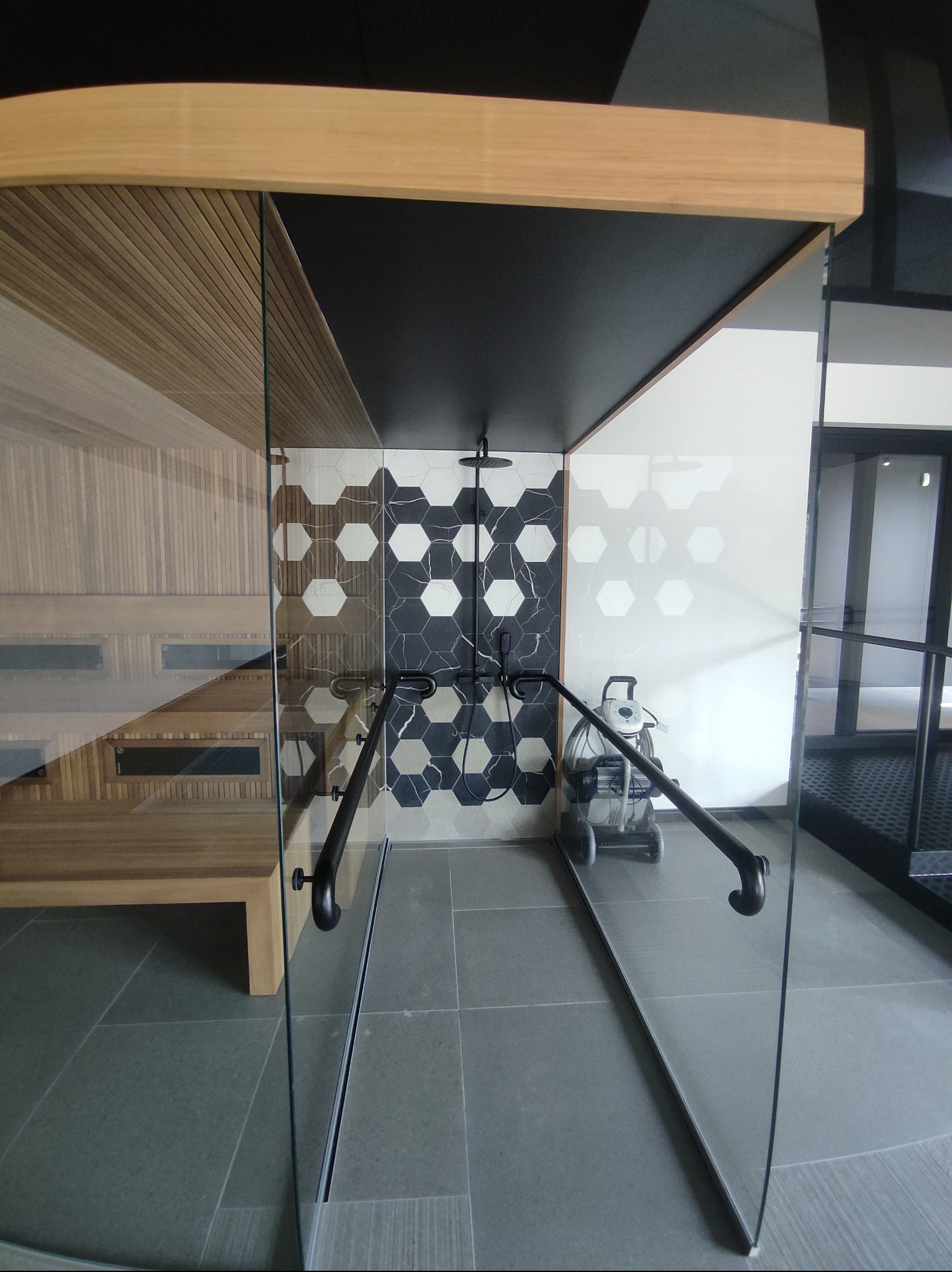 Nowoczesna kabina prysznicowa z czarną armaturą i heksagonalnymi płytkami w odcieniach szarości, widoczna z perspektywy wejścia. Obok sauna z drewnianym wykończeniem.
