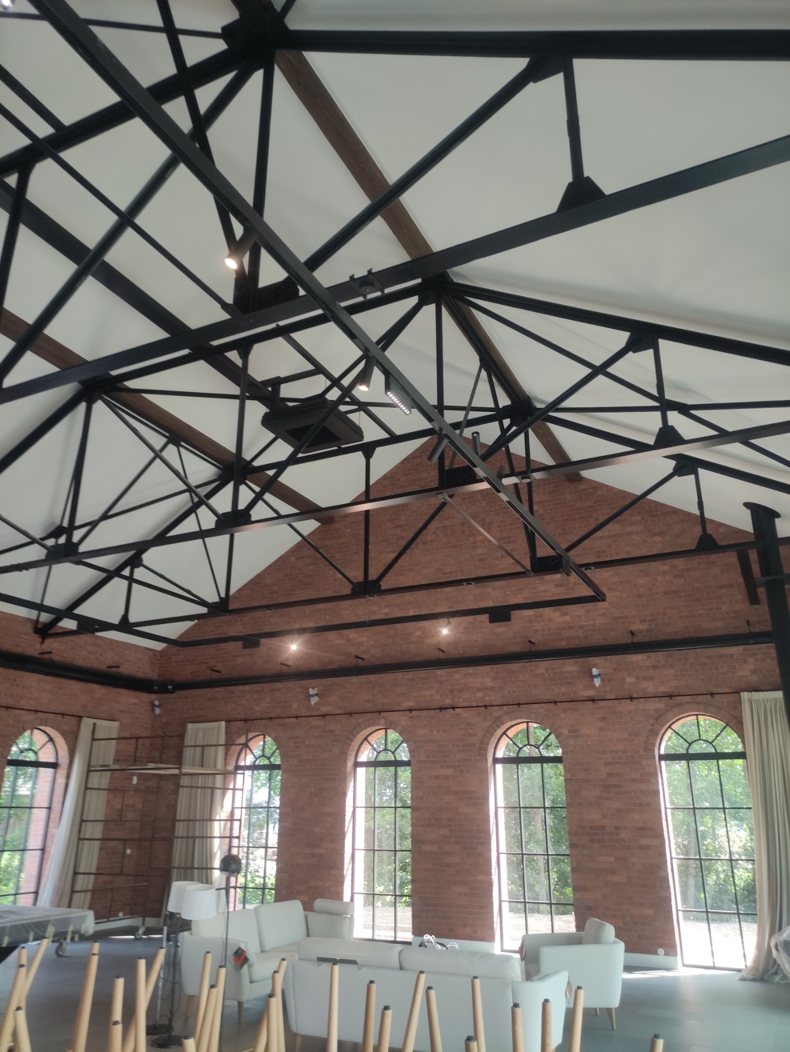 Loftowe wnętrze po remoncie w Bystrej: ceglana ściana, okna łukowe, metalowa konstrukcja dachu i białe meble. Widok na salę z krzesłami i sofami.