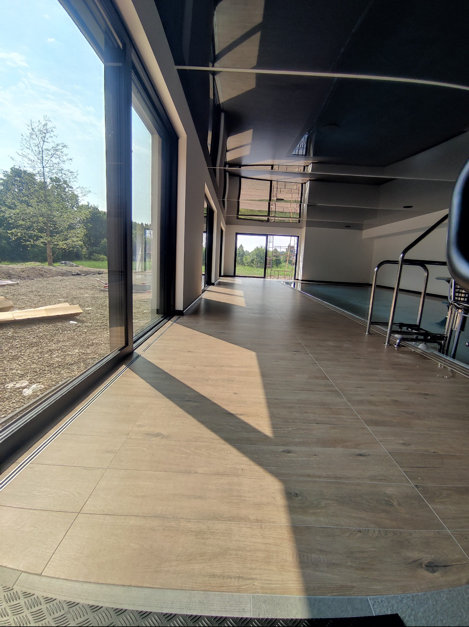 Jasna podłoga z drewnopodobnych płytek w przestronnym wnętrzu z dużymi oknami i czarnym sufitem. Widok na zewnątrz i metalowe schodki.