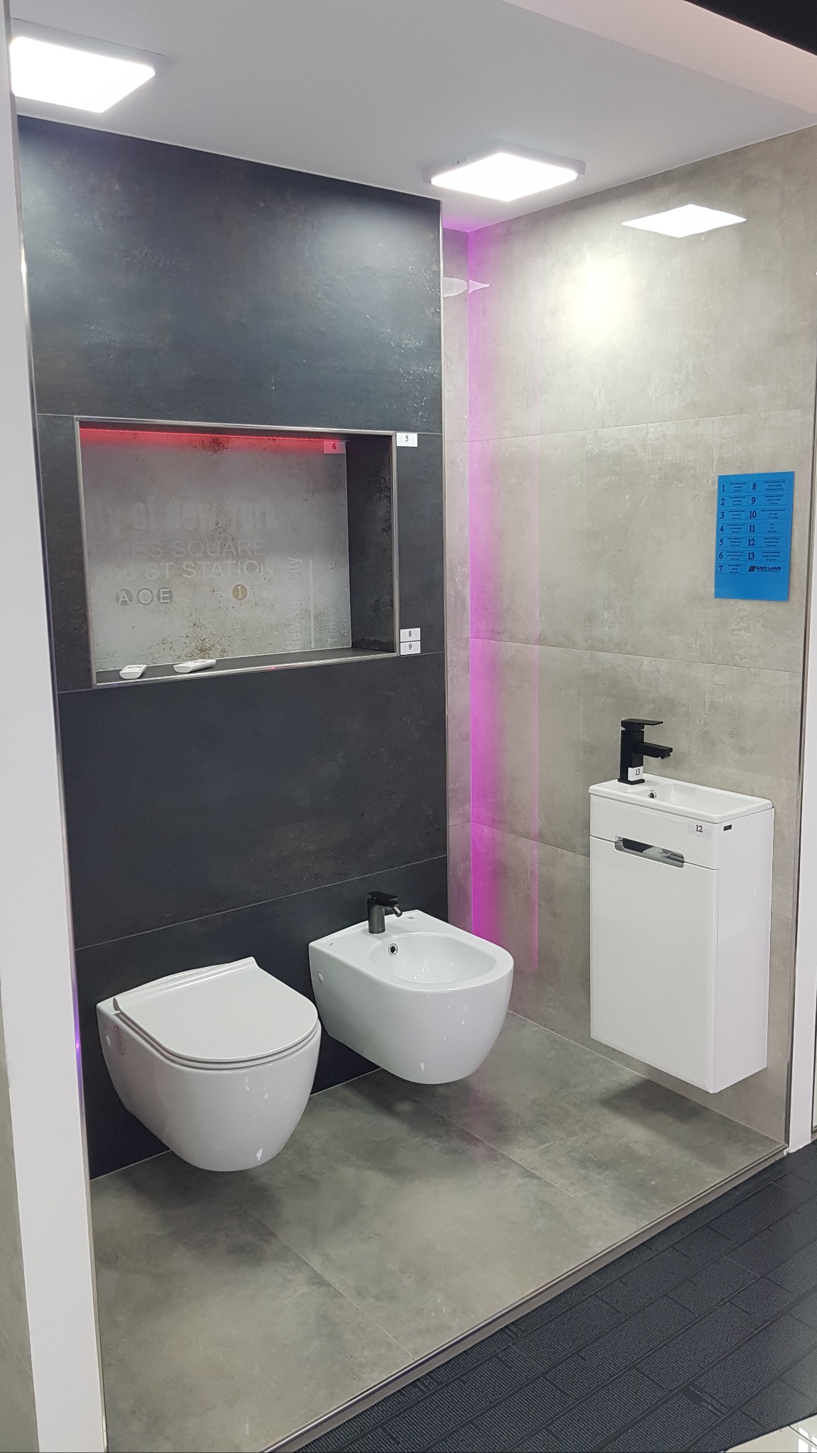 Nowoczesna łazienka z podświetlaną wnęką, umywalką na szafce, toaletą i bidetem. Szare płytki, czarna armatura. Minimalistyczny design, oświetlenie LED.