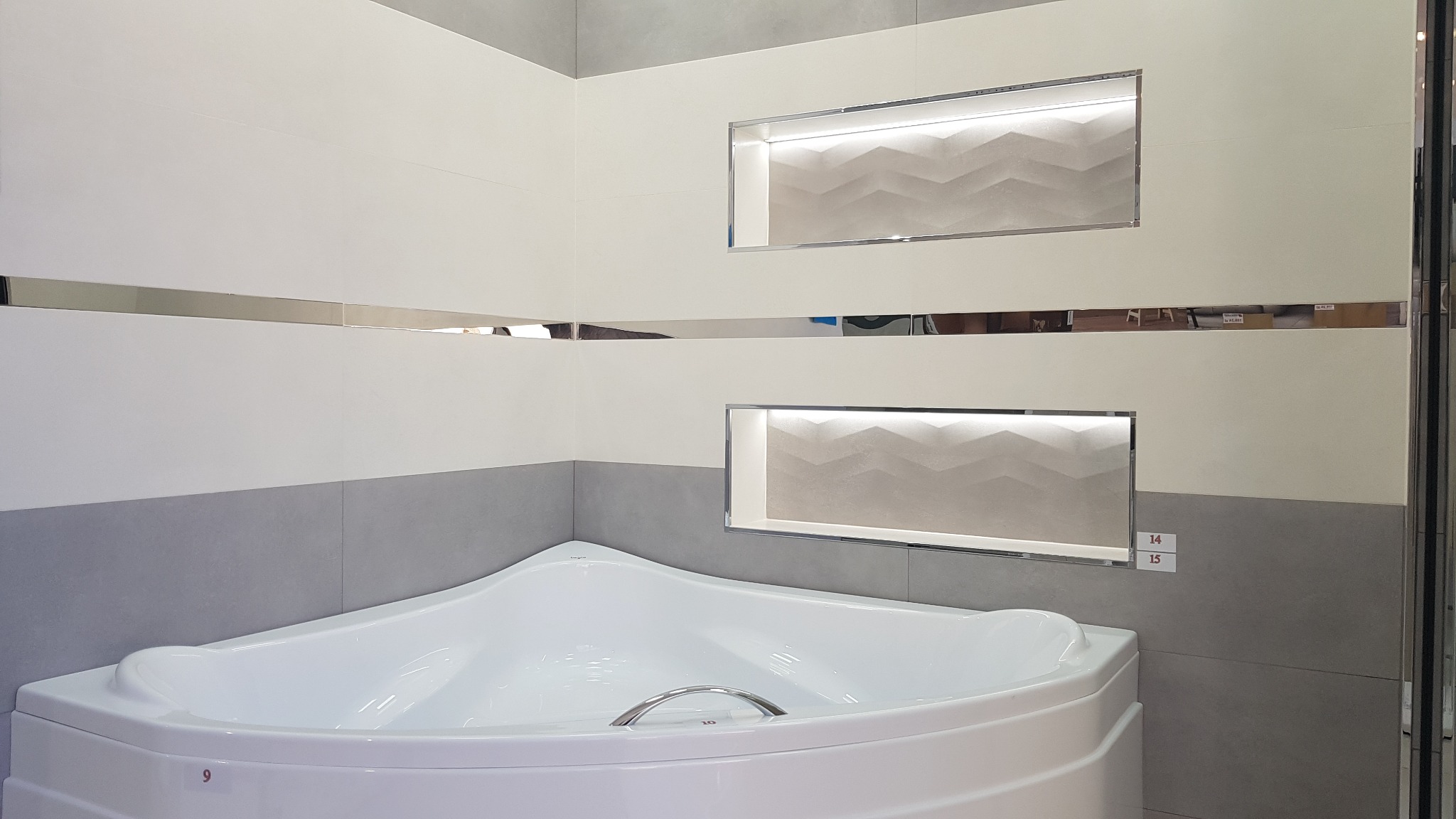 Nowoczesna łazienka z narożną wanną i podświetlanymi wnękami w ścianie wyłożonej biało-szarą glazurą. Chromowane listwy dekoracyjne.