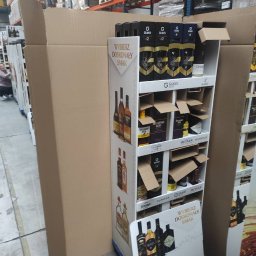 DELTIMA SP. Z O. O. - Wypełniony alkoholem stand reklamowy w magazynie, częściowo zasłonięty kartonem. Widoczne butelki różnych marek alkoholi, regały magazynowe w tle.