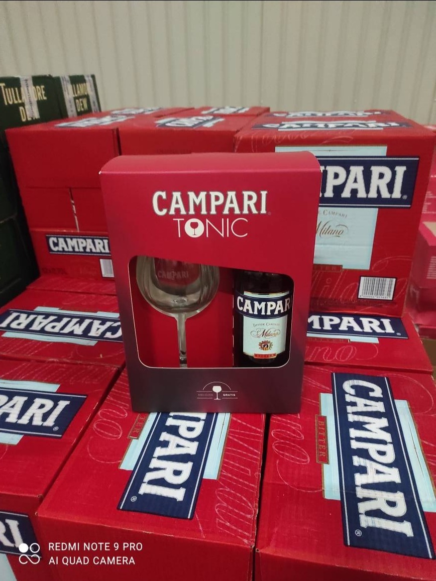 Zestaw Campari Tonic w czerwonym opakowaniu, składający się z butelki, szklanki i wkładki, umieszczony na tle kartonów z logo Campari. Konfekcjonowanie CEDC.