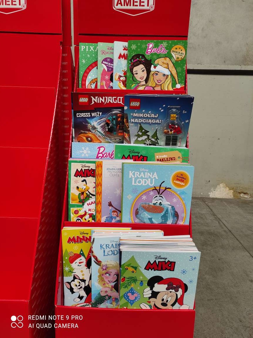 Czerwony stand reklamowy z książeczkami dla dzieci (Barbie, Lego, Kraina Lodu, Miki) na różnych poziomach. Widoczne logo AMEET na górze standu.