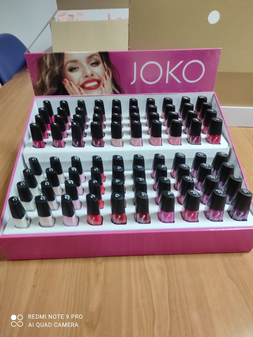 Różowy display z lakierami do paznokci JOKO, ułożonymi w rzędach. W tle kartonowe pudła. Widoczne logo marki i twarz modelki. Konfekcjonowanie kosmetyków.