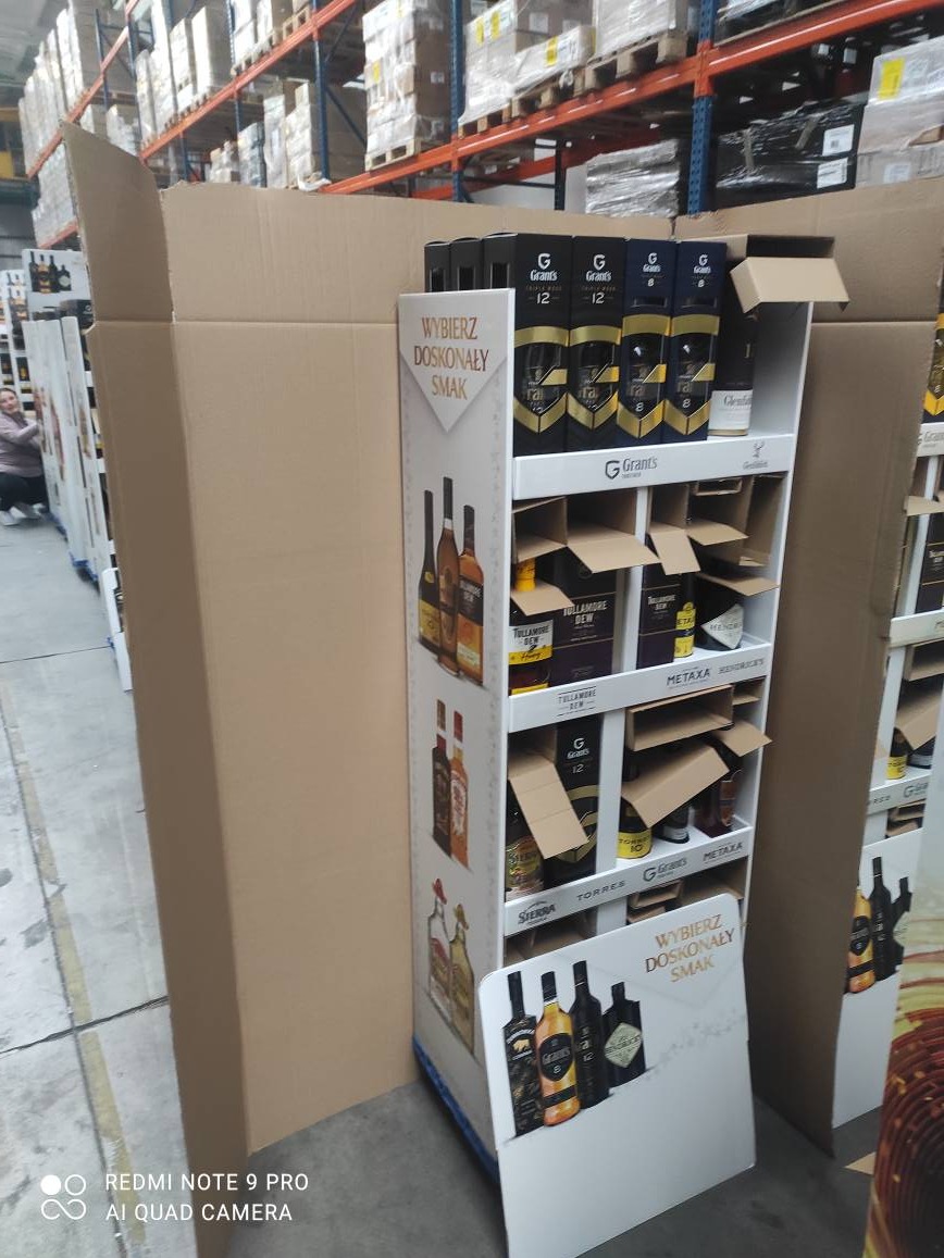 Wypełniony alkoholem stand reklamowy w magazynie, częściowo zasłonięty kartonem. Widoczne butelki różnych marek alkoholi, regały magazynowe w tle.
