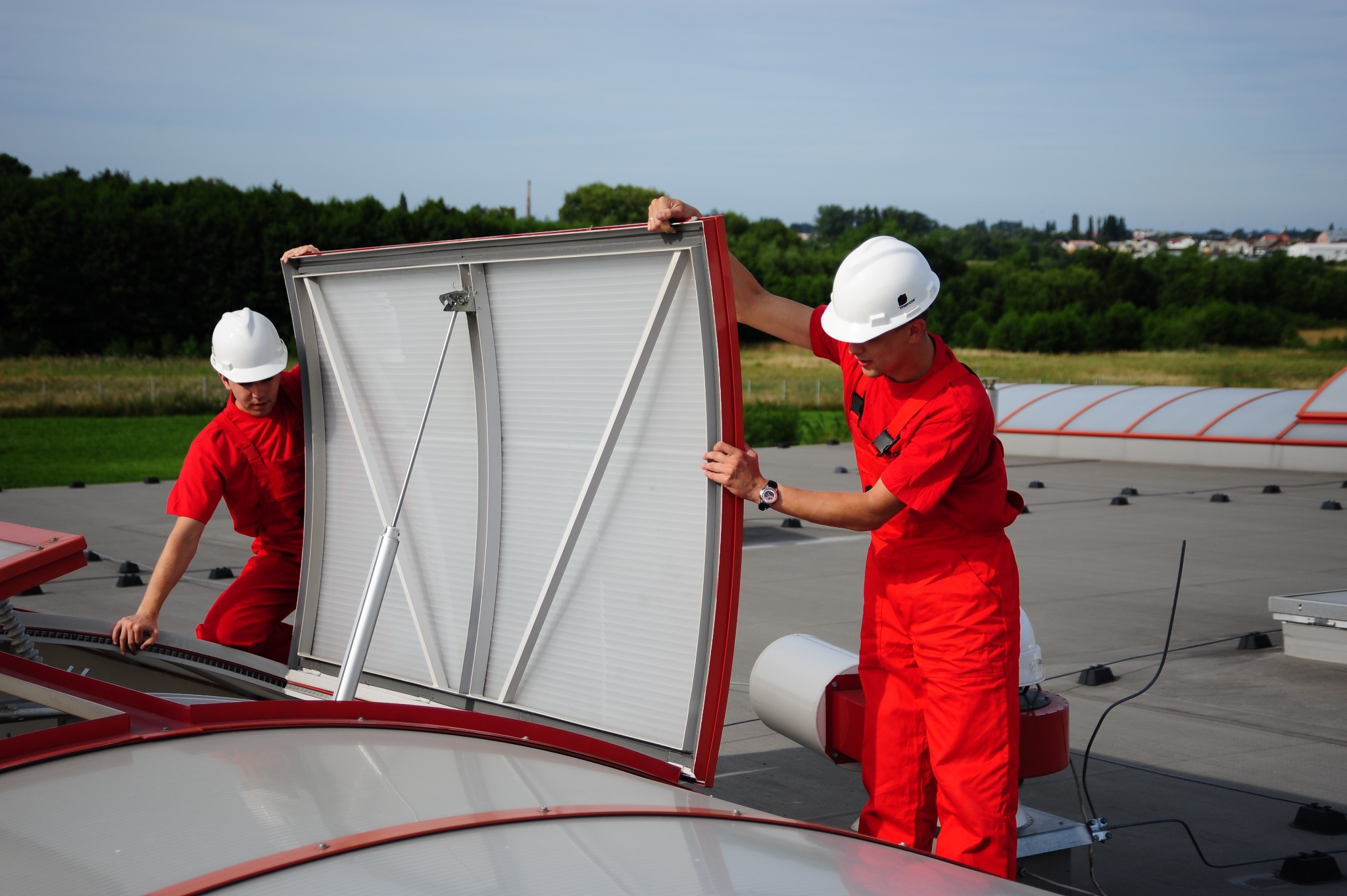 Dwóch pracowników w czerwonych kombinezonach i kaskach BHP otwiera świetlik dachowy na płaskim dachu budynku przemysłowego. Widok na zielone drzewa w tle.