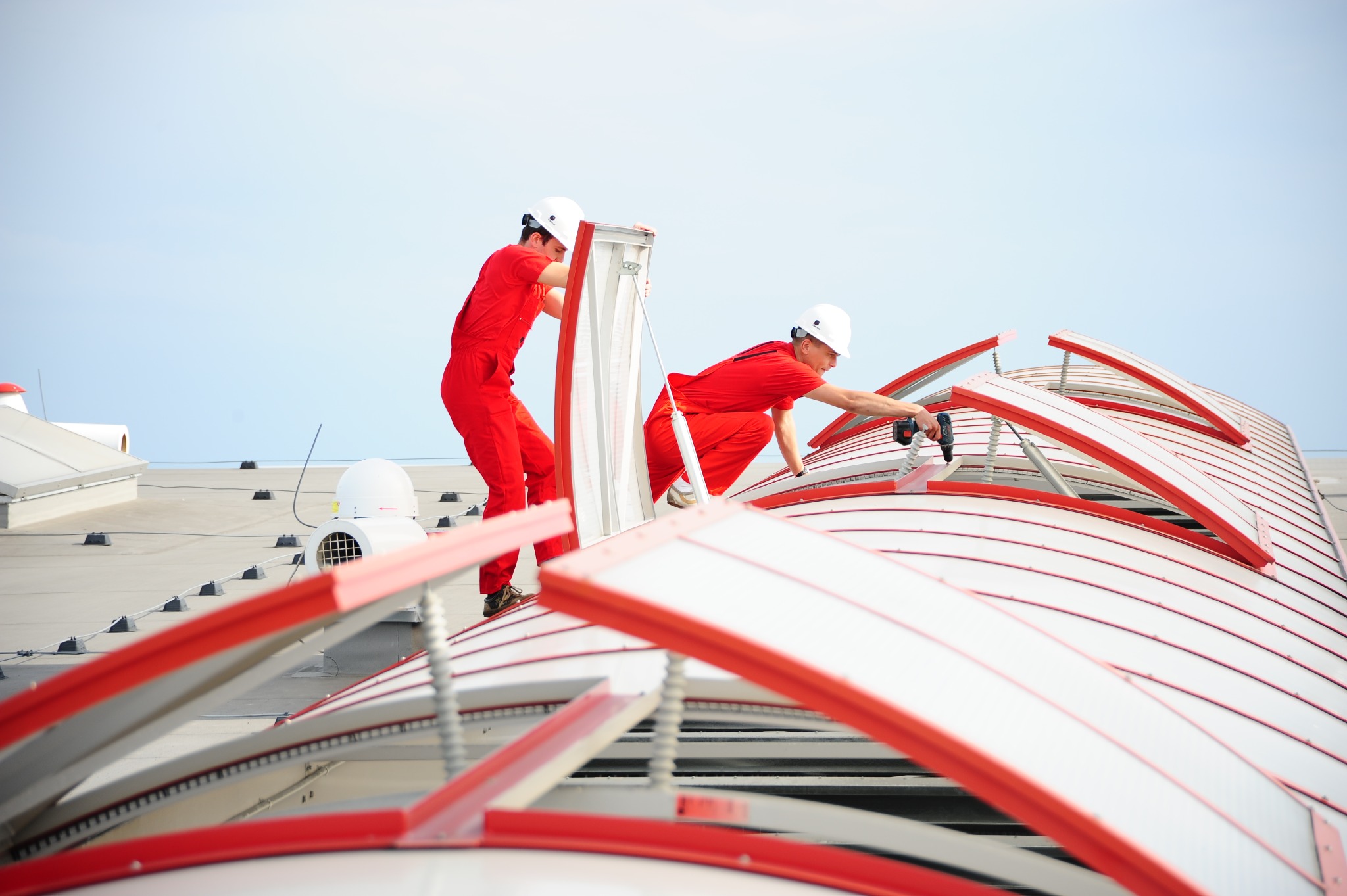 Dwóch monterów w czerwonych kombinezonach i białych kaskach instaluje świetliki dachowe na płaskim dachu budynku przemysłowego przy użyciu wiertarki.
