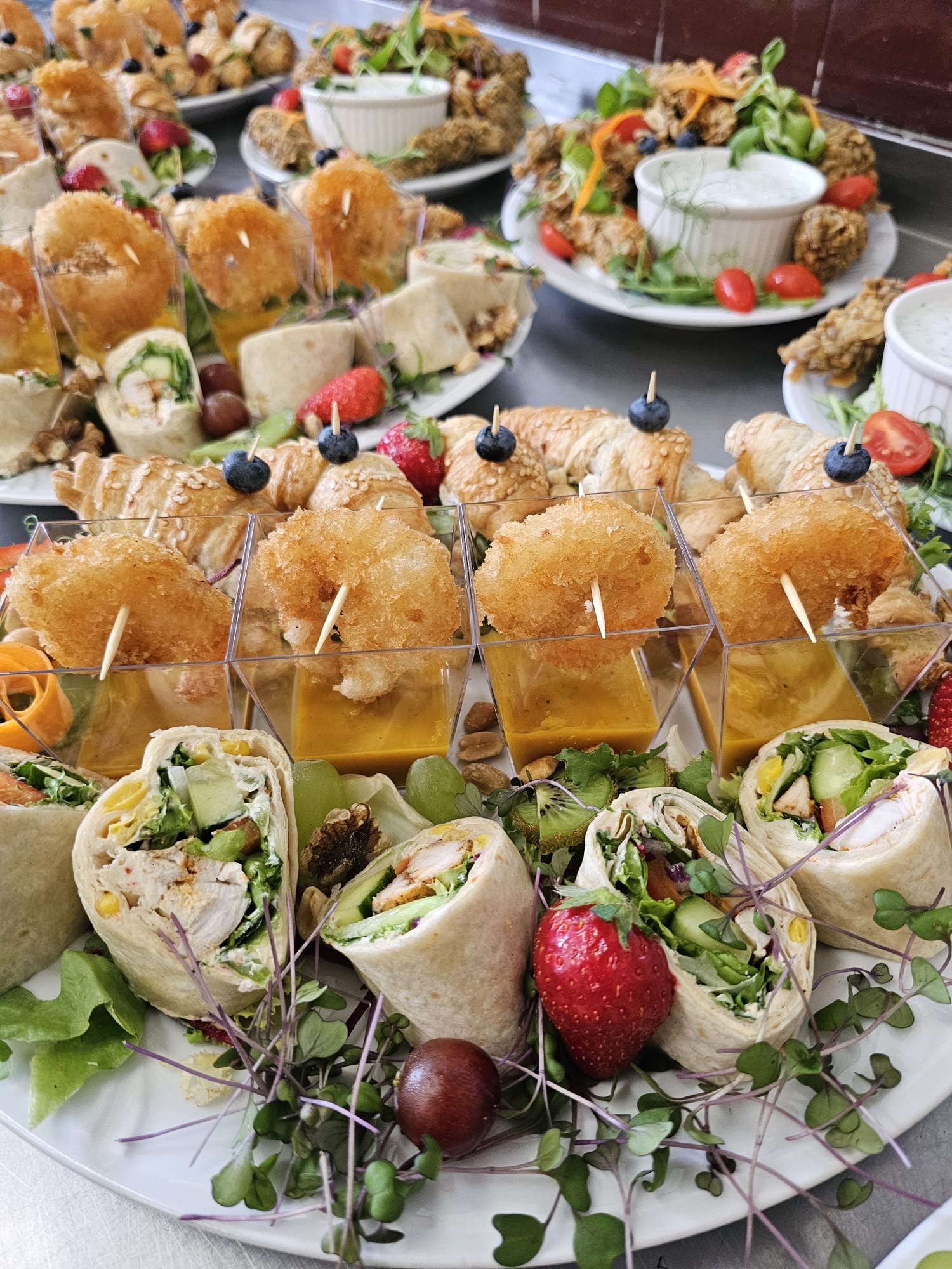 Bogata kompozycja cateringowa: wrap'y z kurczakiem, sałatki, krokiety w szklankach i rogaliki z borówkami na białym talerzu, idealne na firmowe przyjęcie.