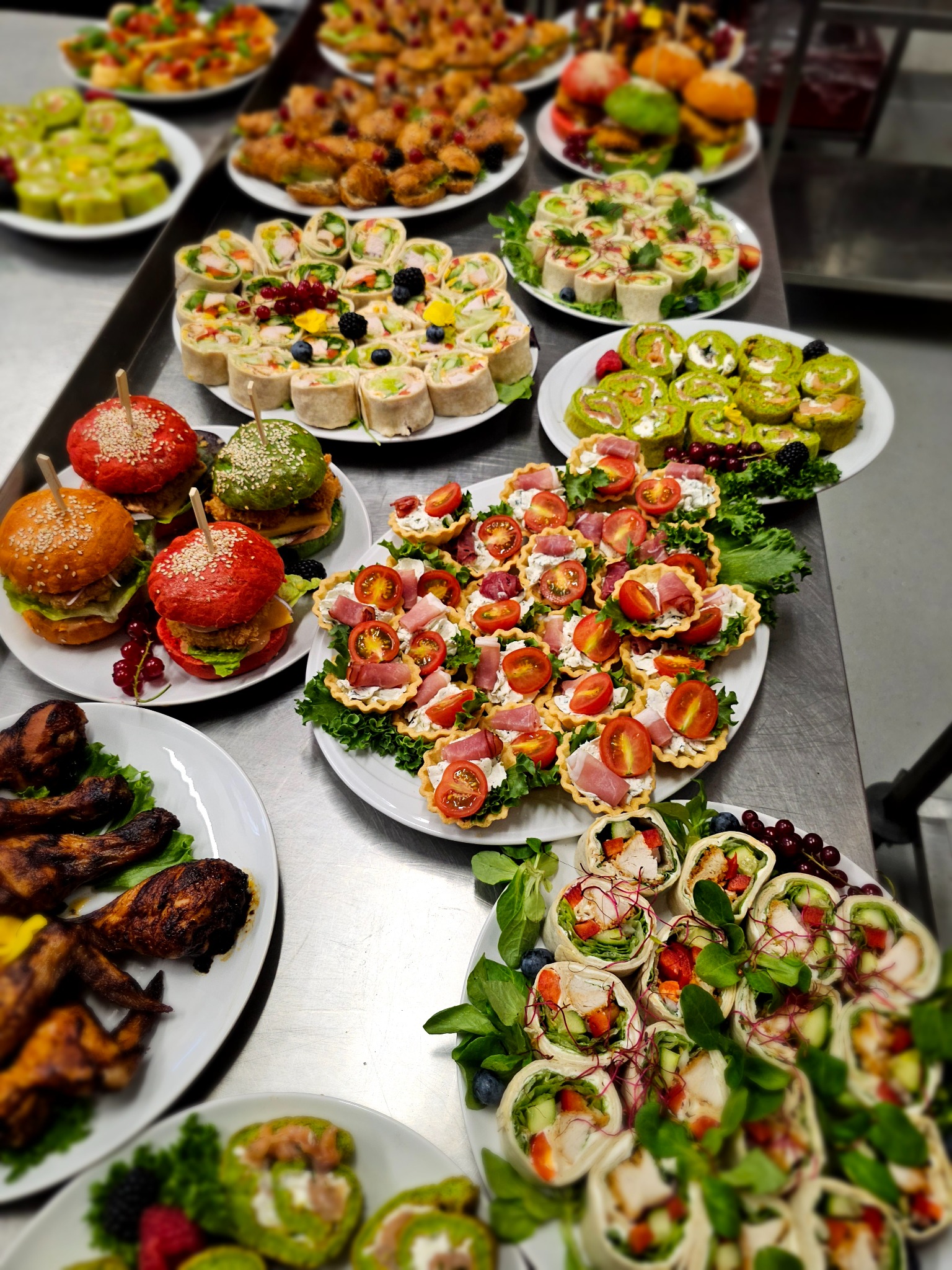 Stół cateringowy z różnorodnymi przekąskami: kolorowe burgery, tartaletki z pomidorami, roladki z warzywami i mięsem, pieczone skrzydełka, dekorowane owocami.