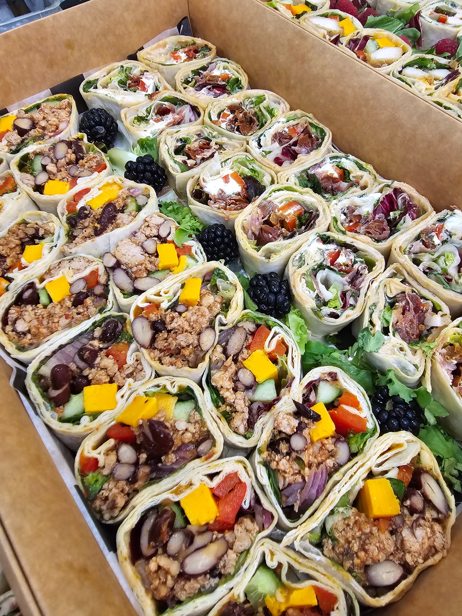 Różnorodny catering: pudełko pełne tortilli z warzywami, mięsem i owocami, ułożonych w rzędach, z dekoracyjnymi jeżynami i sałatą. Zdjęcie z góry.