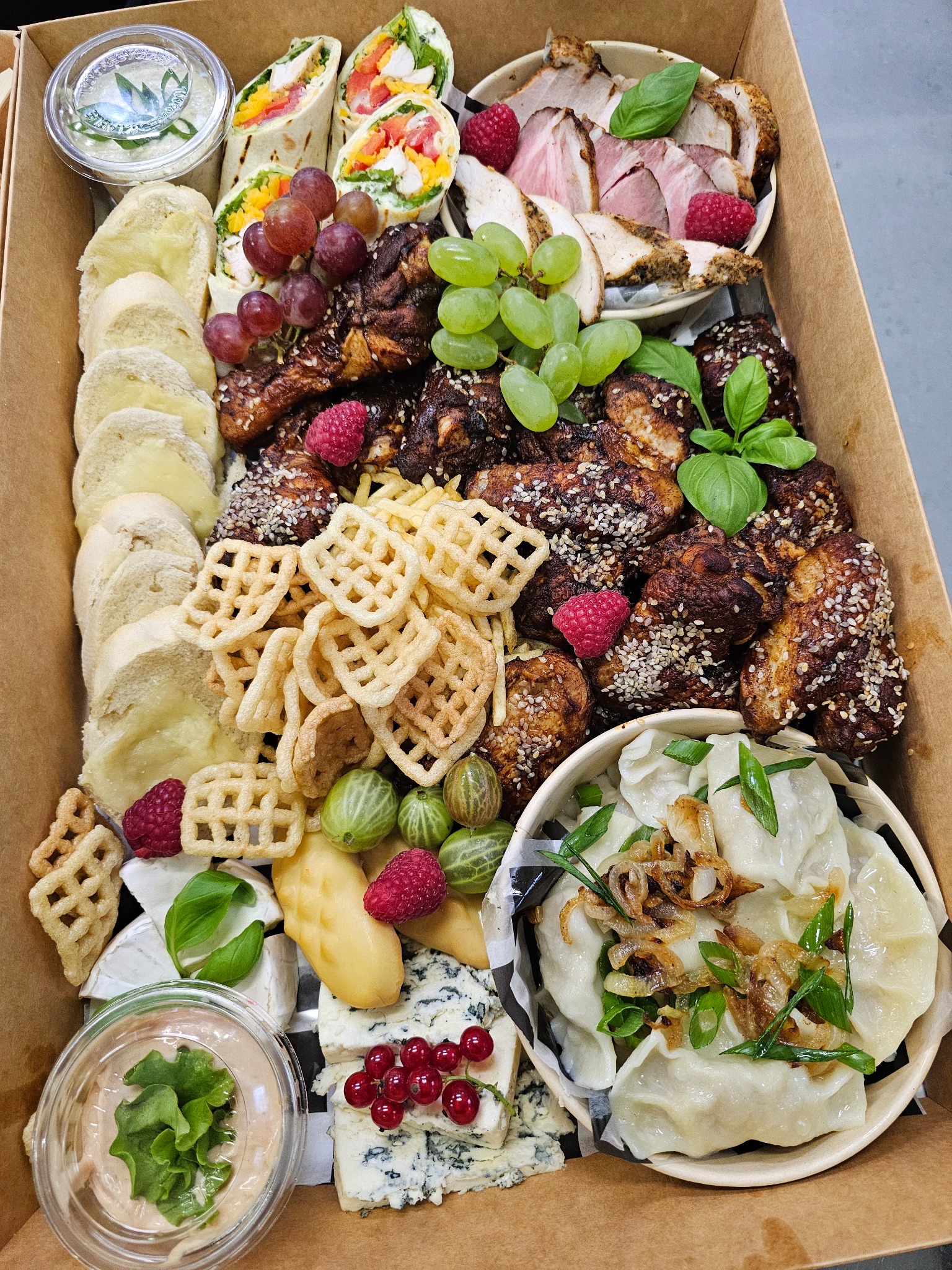 Bogato zaopatrzony karton z cateringiem: roladki, wędliny, sery pleśniowe, pierogi z cebulką, owoce i sosy. Różnorodność smaków i kolorów, idealne na przyjęcie.