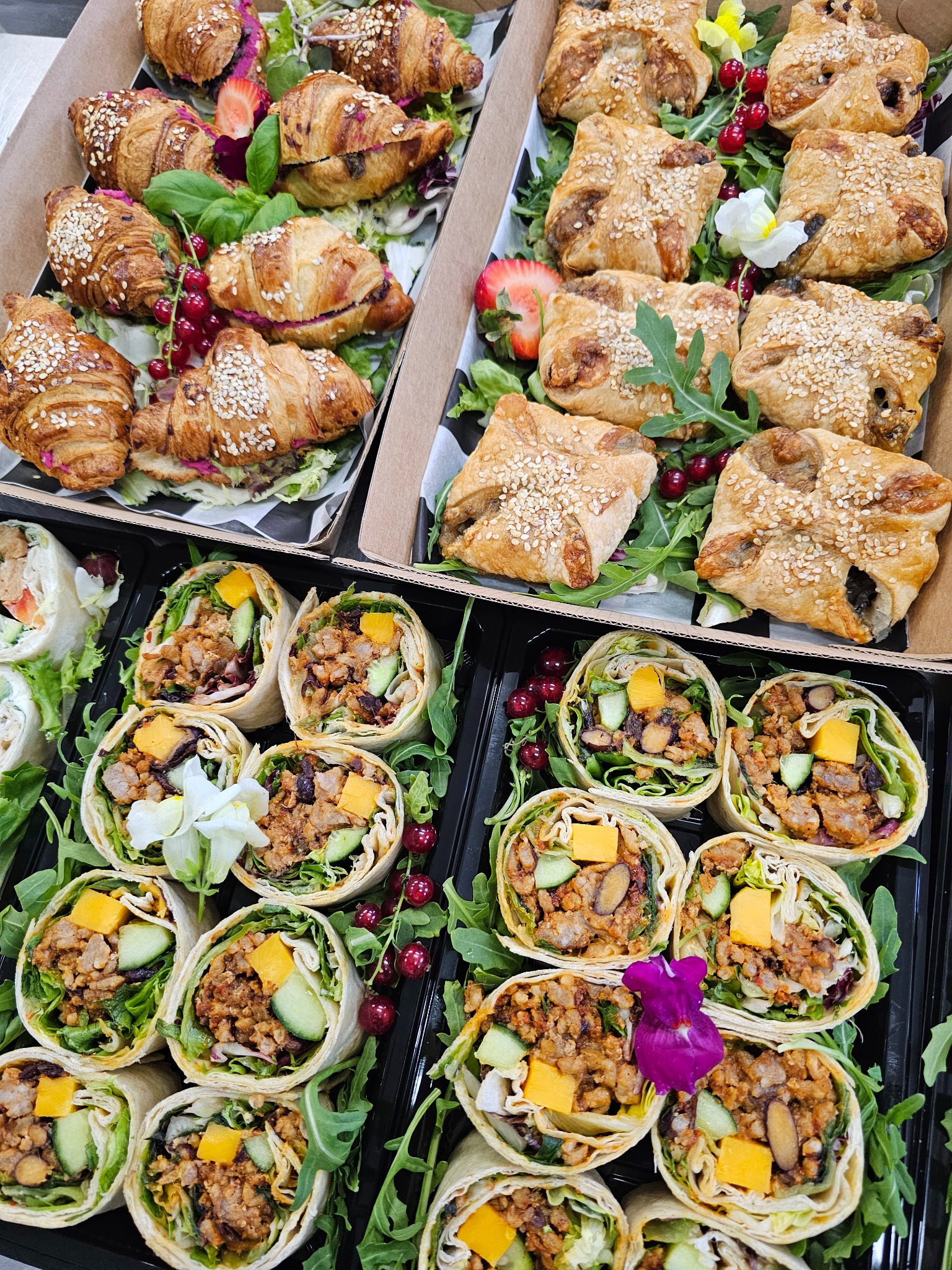 Elegancki catering: rogaliki z sezamem, kwadratowe ciastka i tortille z warzywami, ułożone na rukoli z dekoracją z kwiatów i owoców. Gotowe do serwowania.