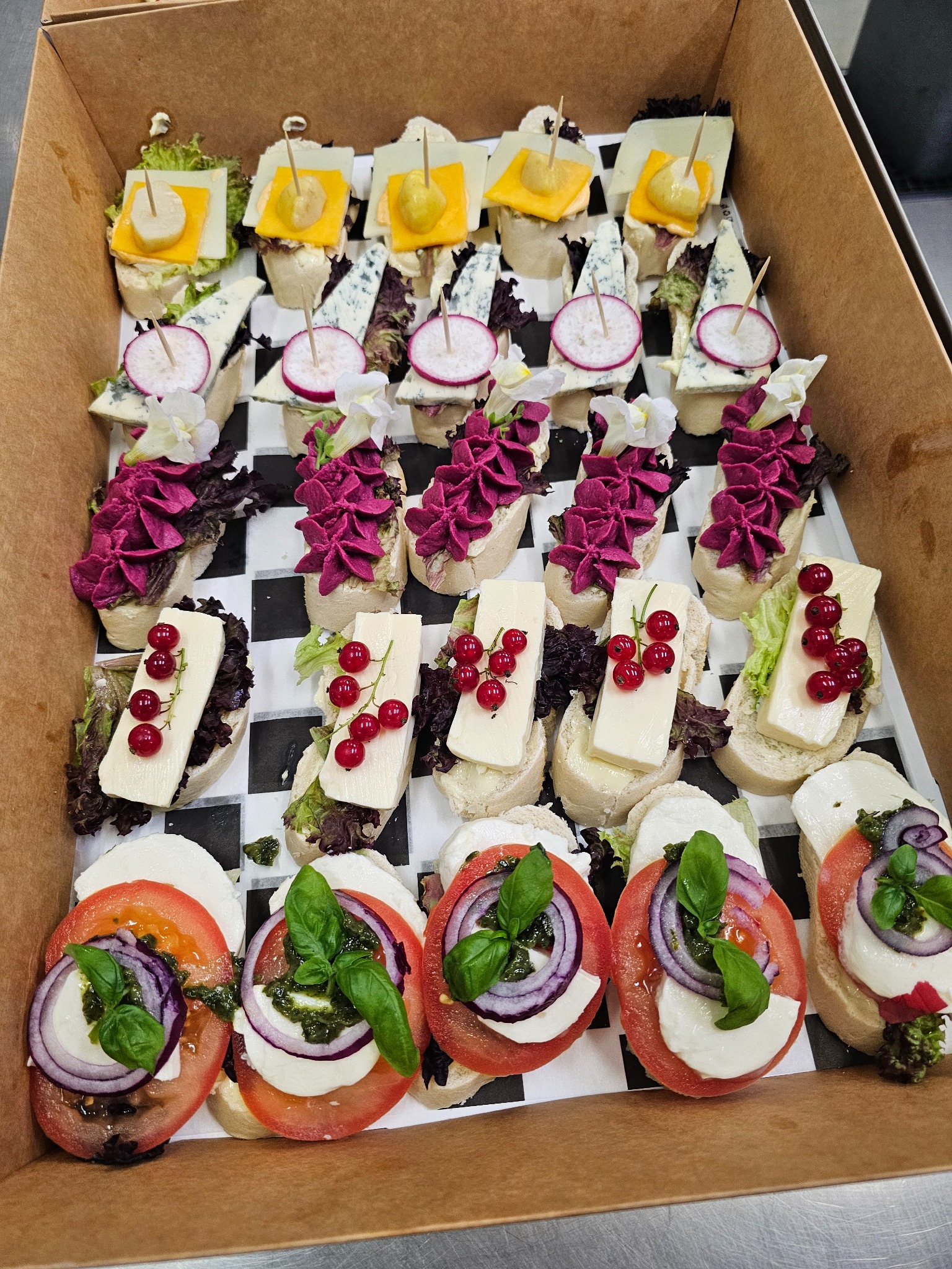 Różnorodne kanapeczki na pieczywie w kartonowym pudełku: ser żółty, pleśniowy, mozzarella z pomidorem i bazylią, dekorowane rzodkiewką, kwiatami i czerwoną porzeczką.