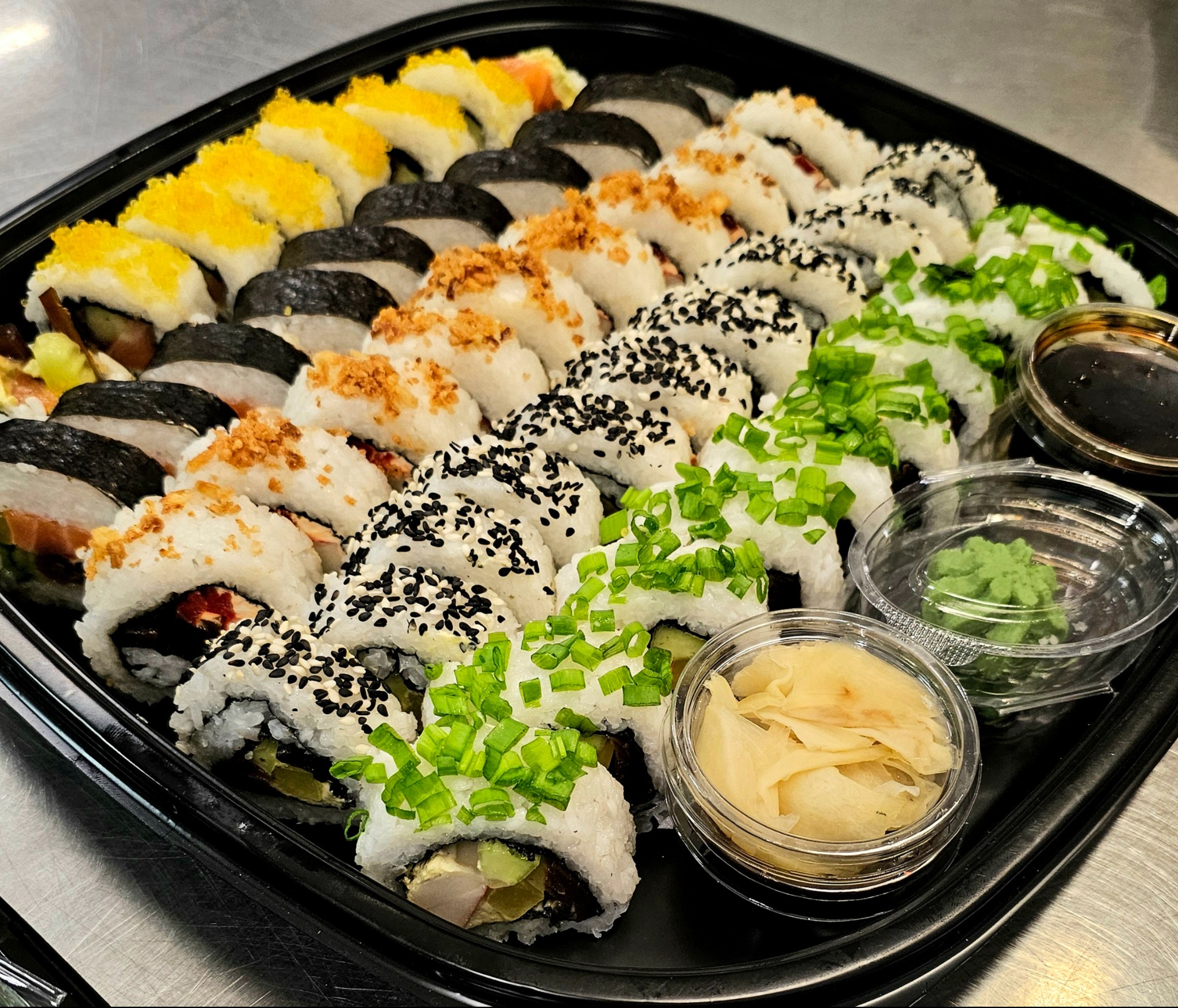 Zestaw sushi z różnymi dodatkami, w tym kawior, sezam i szczypiorek, ułożony na czarnej tacy z sosem sojowym, imbirem i wasabi. Blat ze stali nierdzewnej w tle.
