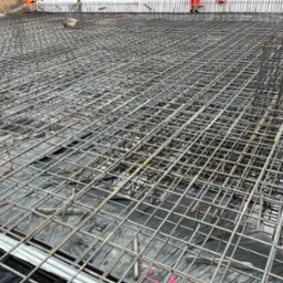 Siatka zbrojeniowa na czarnej folii, przygotowana do zalania betonem. Na obrzeżach widoczne fragmenty szalunków i pracownicy w pomarańczowych kombinezonach.