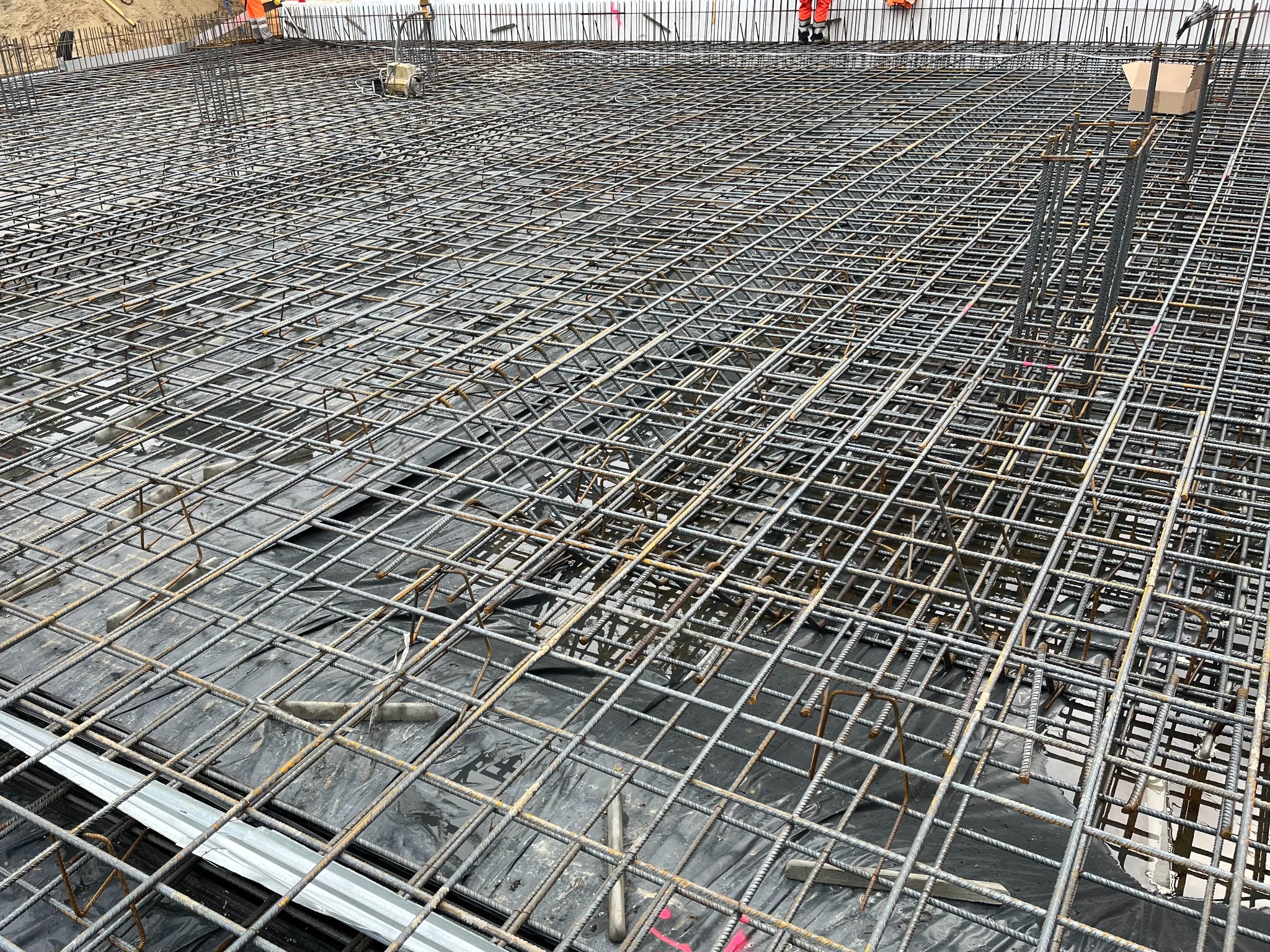 Siatka zbrojeniowa na czarnej folii, przygotowana do zalania betonem. Na obrzeżach widoczne fragmenty szalunków i pracownicy w pomarańczowych kombinezonach.