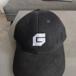 GRUPA CMYKI KATARZYNA PANKIEWICZ - Czarna czapka z daszkiem z haftowanym, białym logo w kształcie litery 'G' na tle szarej kanapy. Widoczne detale szwów i faktury materiału.