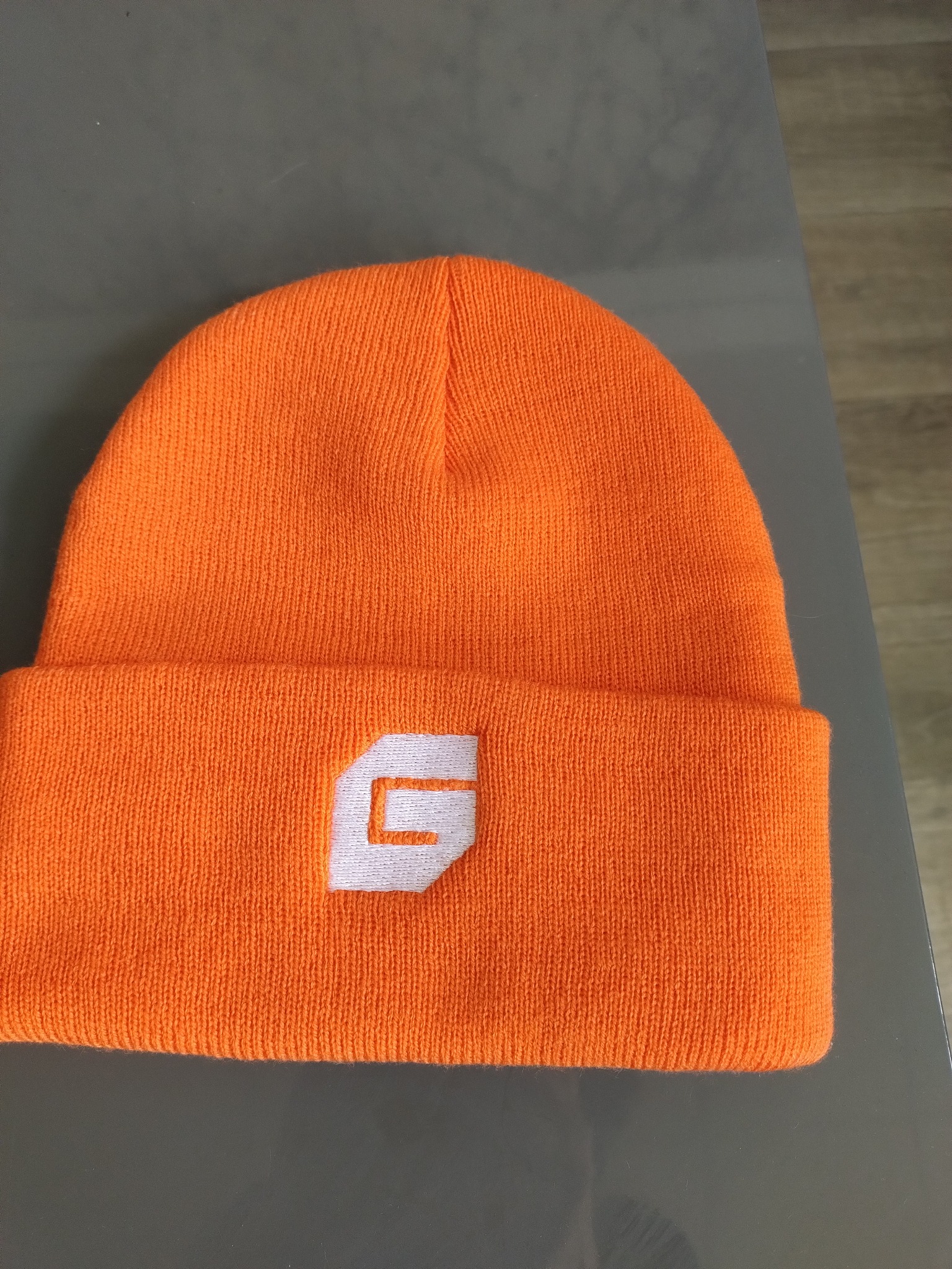 Pomarańczowa czapka zimowa z białym, stylizowanym logo na wywinięciu, leżąca na szarym tle. Widoczna faktura dzianiny.