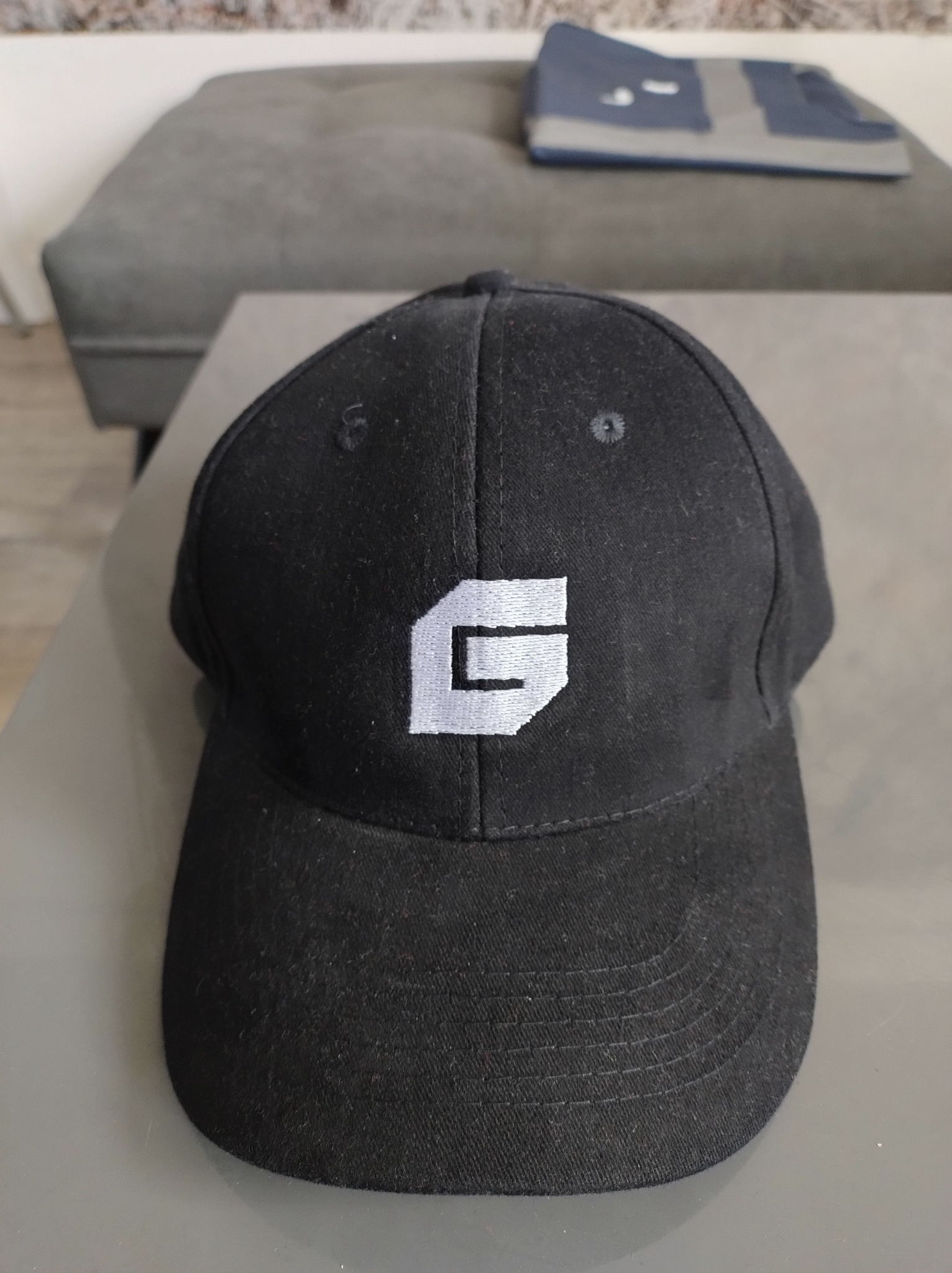 Czarna czapka z daszkiem z haftowanym, białym logo w kształcie litery 'G' na tle szarej kanapy. Widoczne detale szwów i faktury materiału.