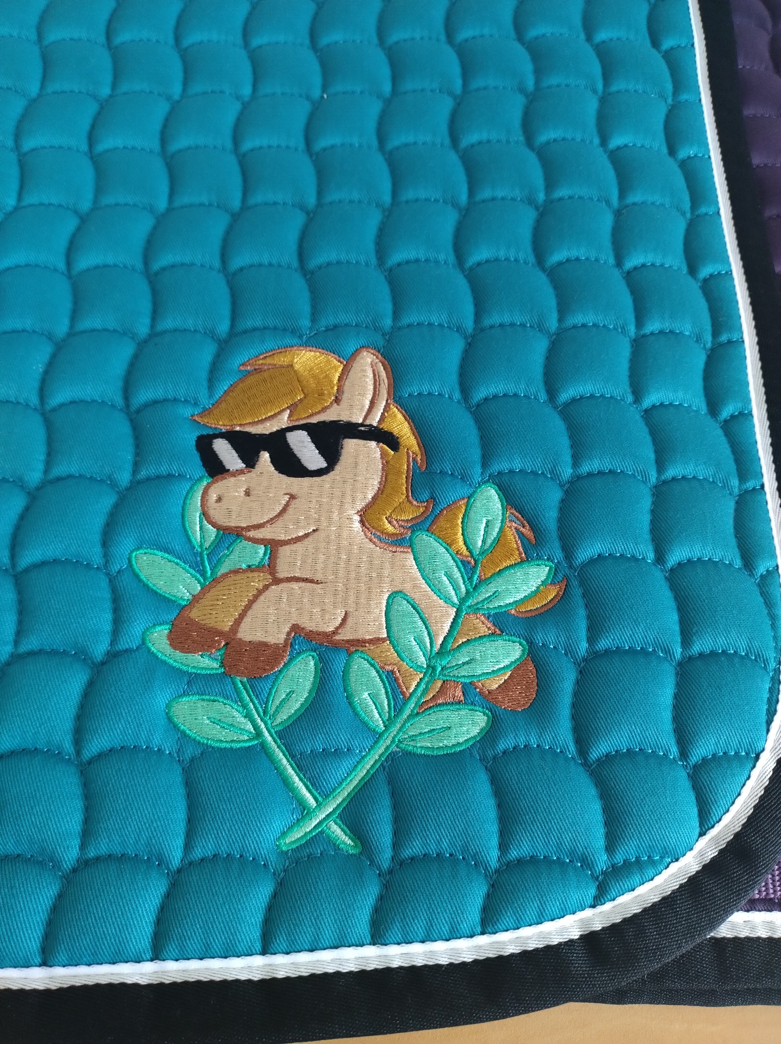 Turkusowa podkładka pod siodło z haftowanym, uśmiechniętym konikiem w okularach przeciwsłonecznych i zielonych liściach. Pikowana powierzchnia, białe obszycie.