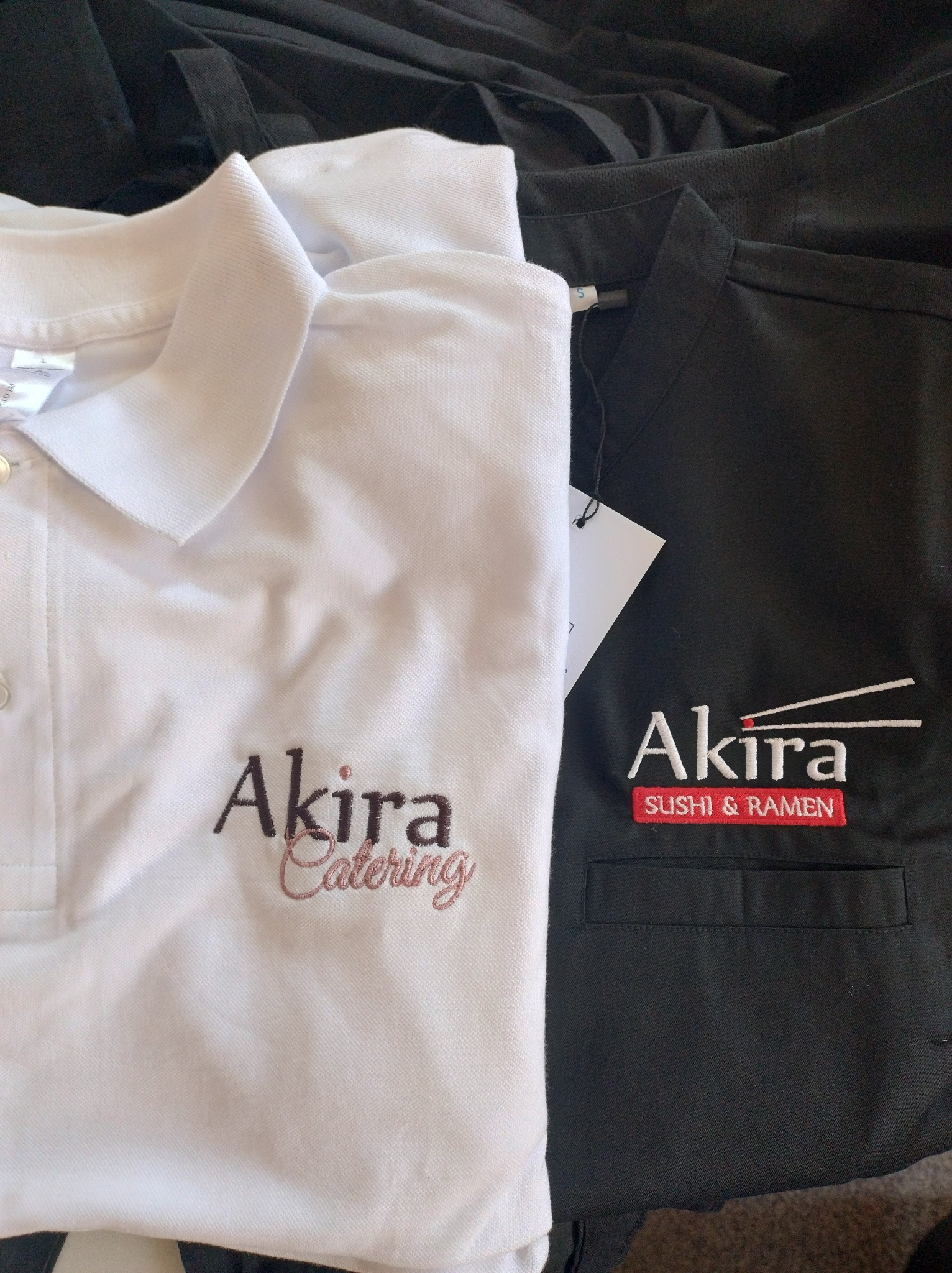 Biała koszulka polo i czarna bluza robocza z wyhaftowanym logo 'Akira', prezentujące usługi haftowania dla branży gastronomicznej.